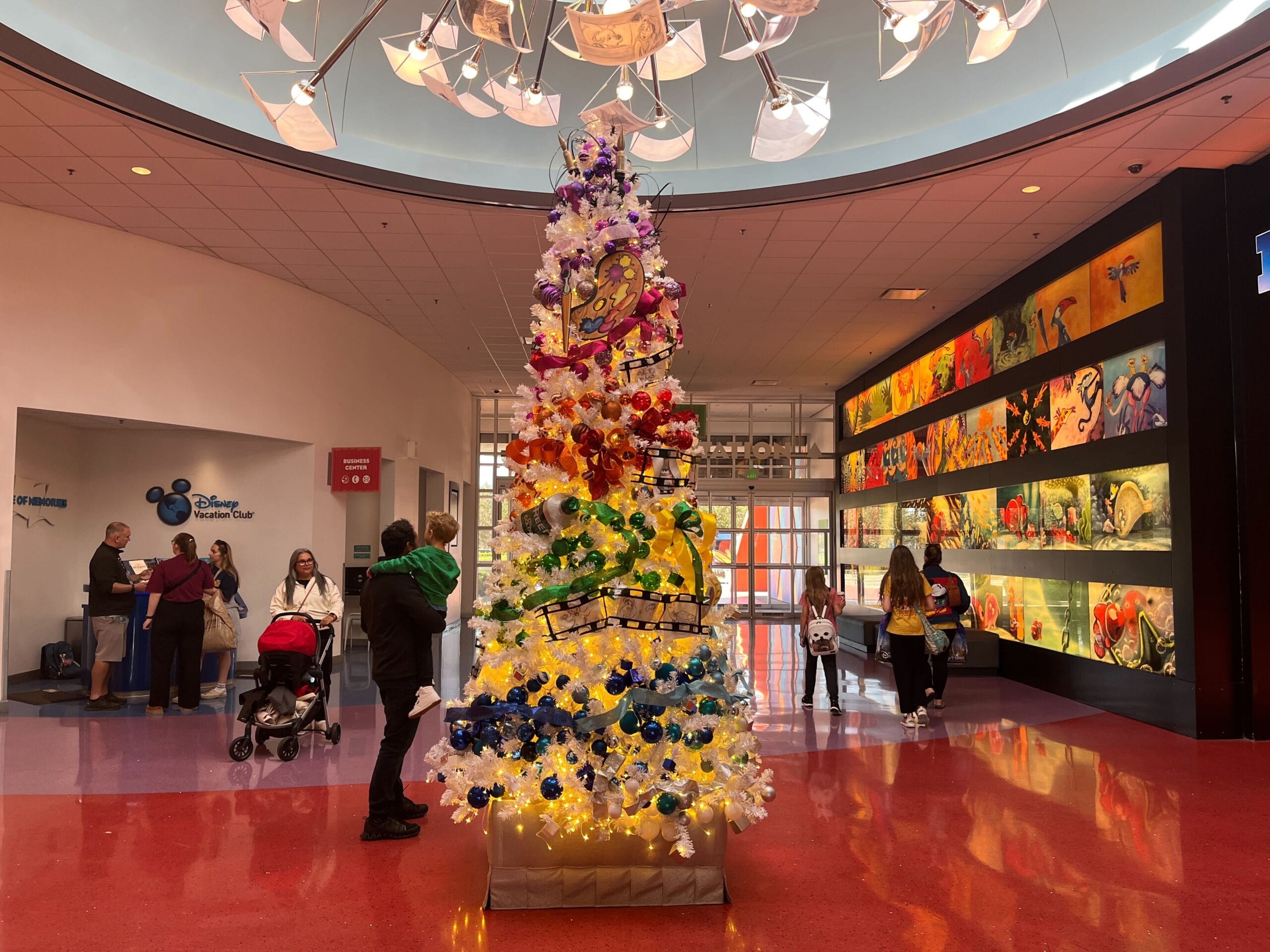 Colorful Christmas tree with decorations stands in a lobby area, surrounded by people. Bright murals line the walls, and a sign reads Disney Photopass Service.