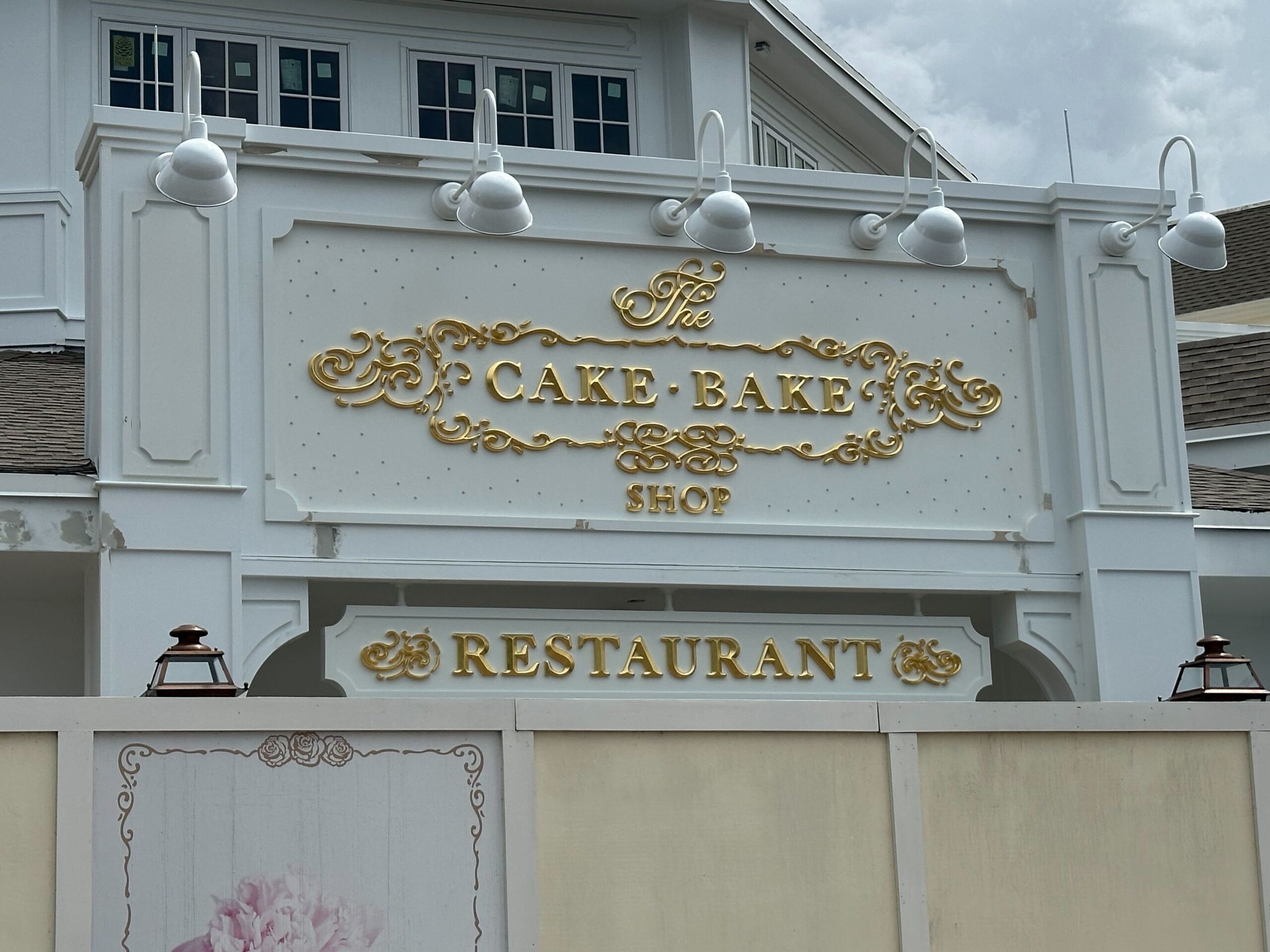 A white building with an ornate gold sign reading The Cake Bake Shop and another sign below it reading Restaurant. Four light fixtures are mounted above the top sign.