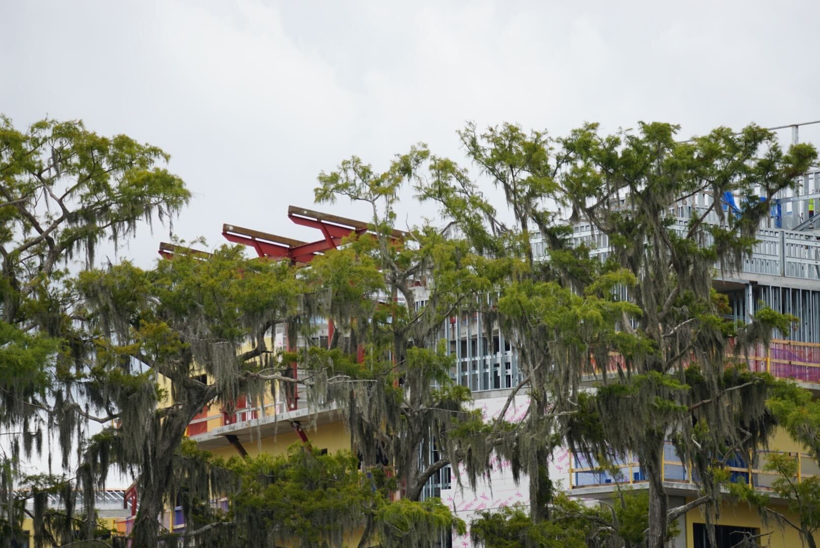 Trees draped with moss partly obscure a colorful Lakeshore building with balconies and red beams under a cloudy sky.