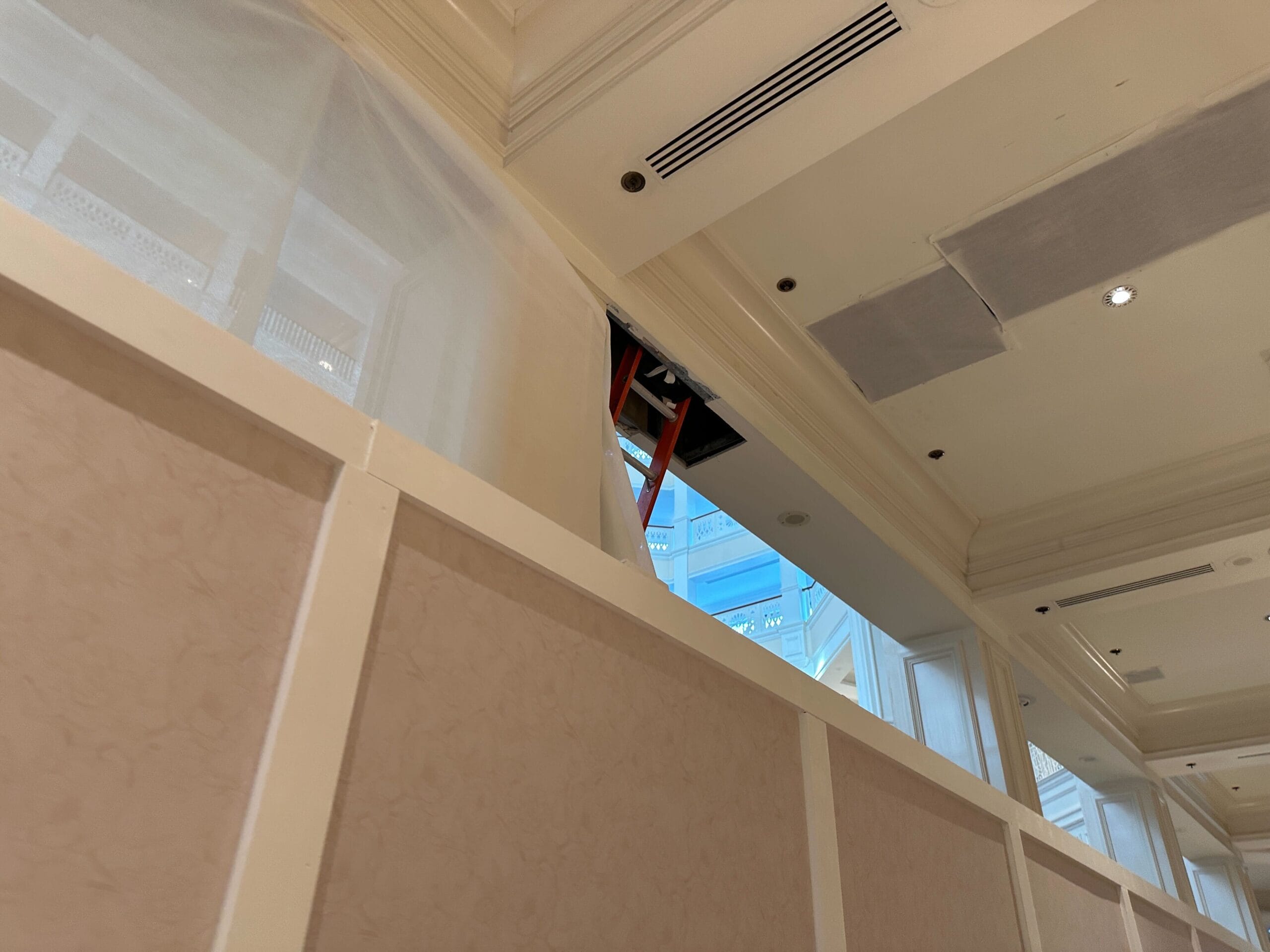 Ceiling panels are open with an orange ladder leaning into the ceiling above a beige partition wall in an indoor space.