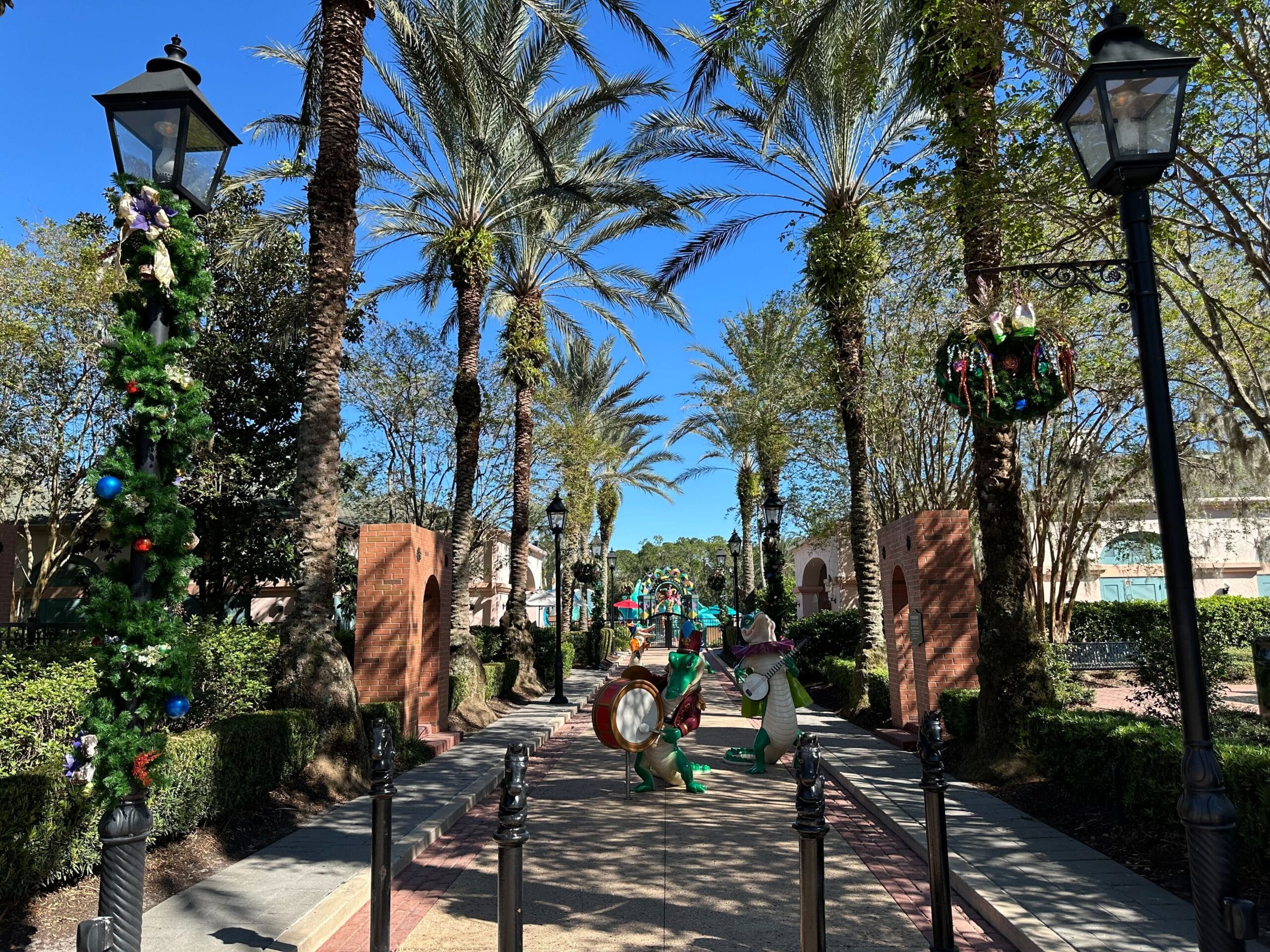 A scenic pathway with tall palm trees, festive green and red decorations, and large decorative characters in a sunny park setting.