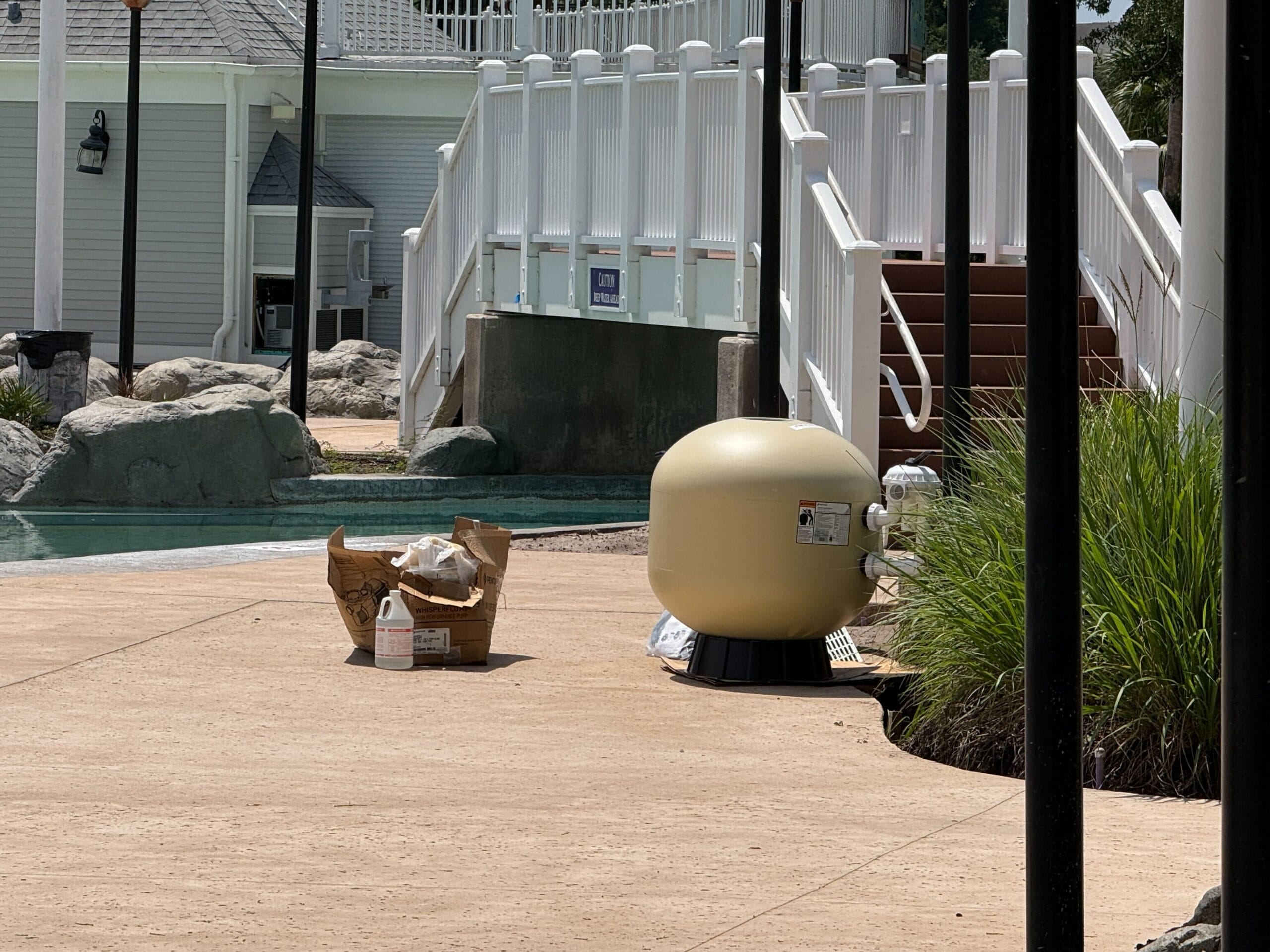A tan pool filter sits on a patio near cleaning supplies, next to a pool and white railing on a sunny day.