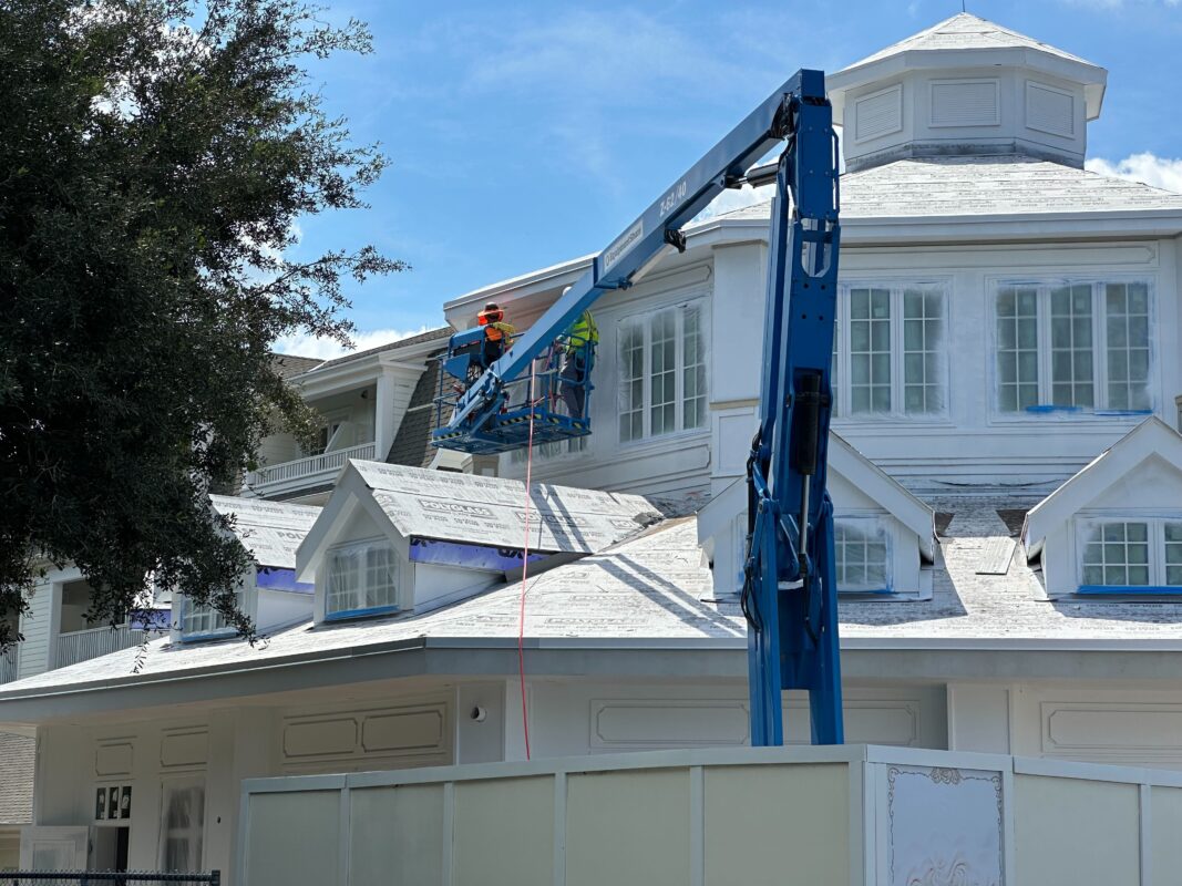 Two workers in safety gear are on a raised platform working on the roof of a large white building on a sunny day.