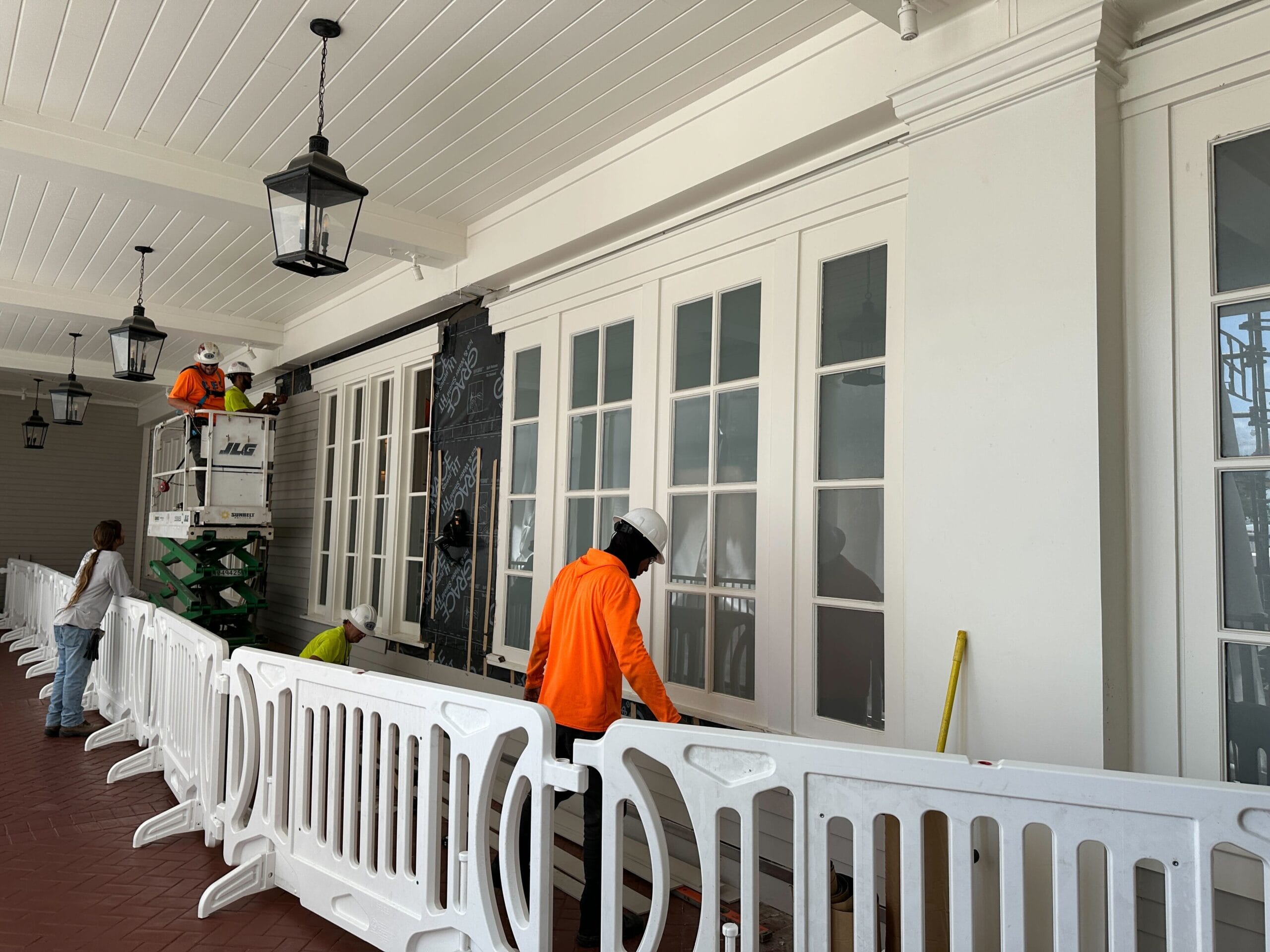 Workers in orange vests repair or paint a white building behind safety barriers indoors.