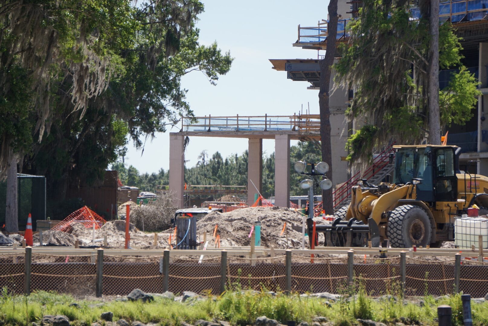 Construction site with machinery, dirt piles, and partially built structures surrounded by trees and fencing.