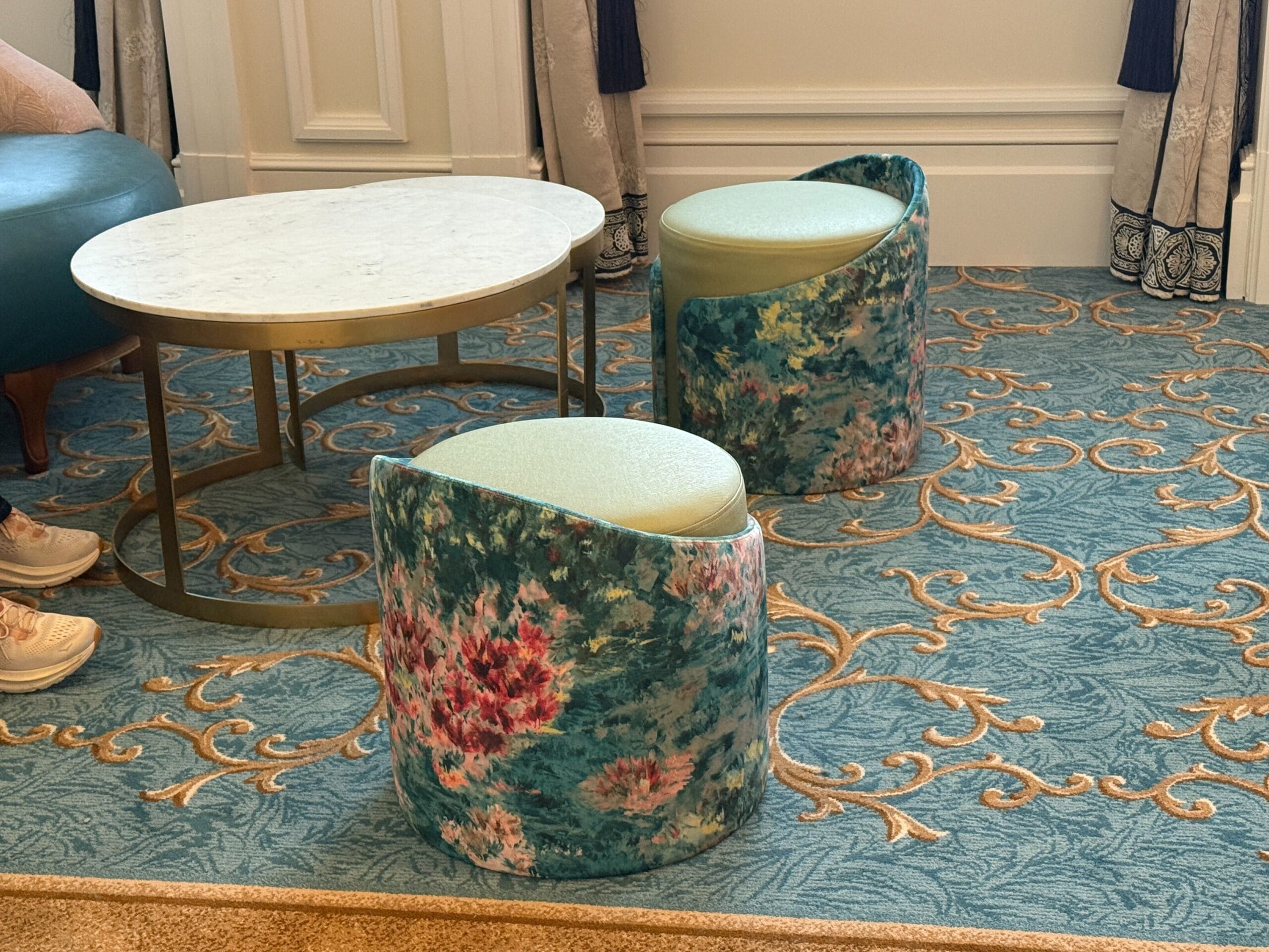 Two floral upholstered chairs and a round marble-top table on an ornate blue and gold carpet. Sneakers visible on the left.