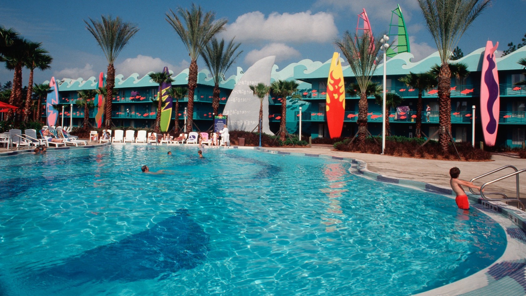 Outdoor swimming pool surrounded by loungers, palm trees, and colorful surfboard decor, with several people swimming and relaxing. The multi-story building with a wave-like design in the background gives off an All-Star resort vibe, perfect for a sporty getaway.
