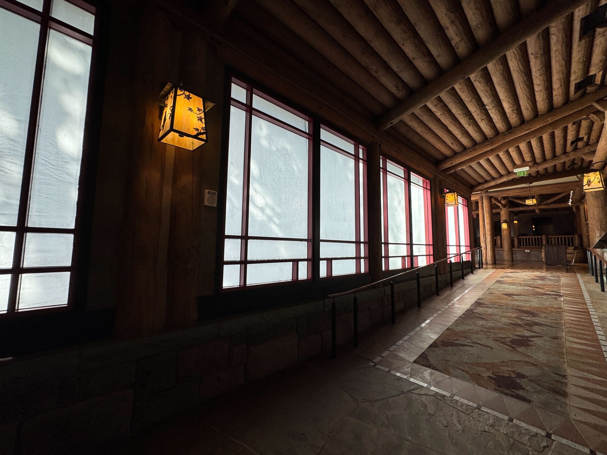 Dimly lit hallway with log ceiling, lanterns, stone floor, and large frosted windows along one side.