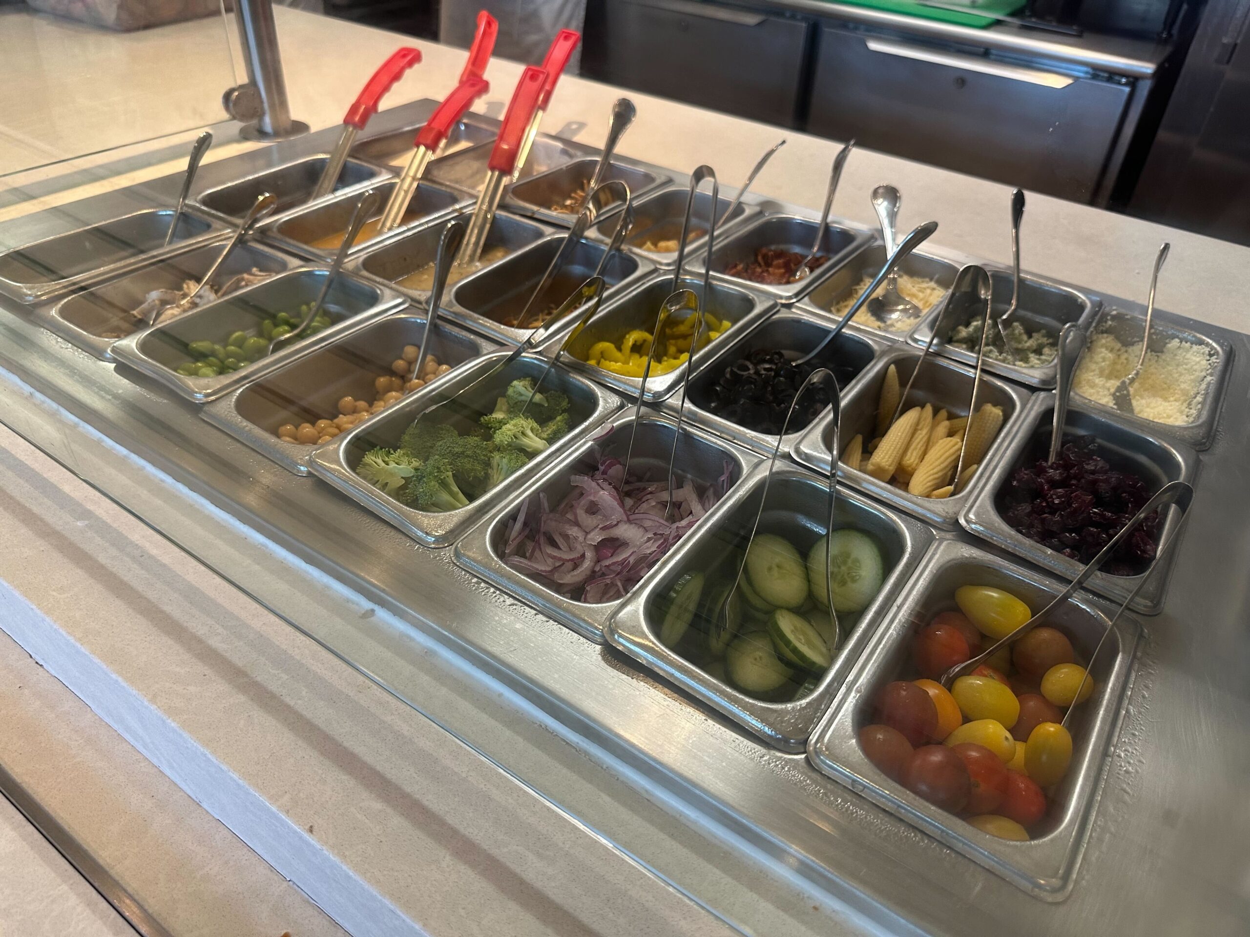 A salad bar with various vegetables and toppings, including cherry tomatoes, cucumber slices, red onions, broccoli florets, chickpeas, olives, and corn arranged in metal containers with serving tongs.