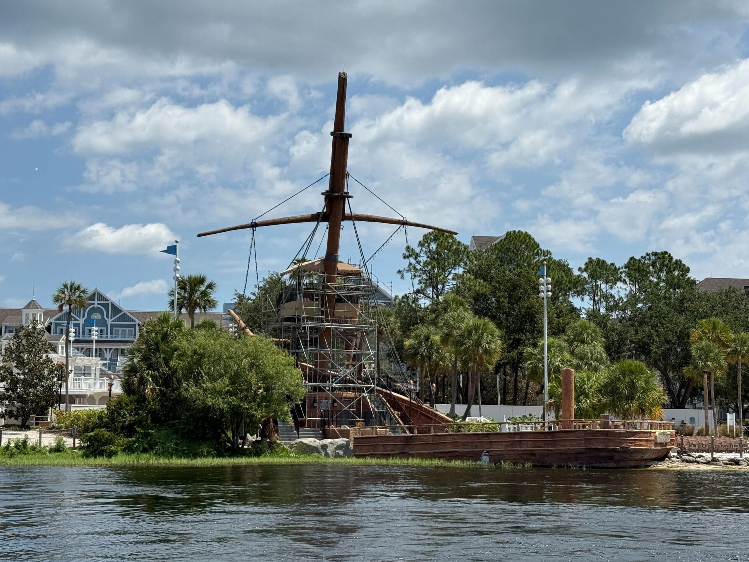 A wooden ship under construction or renovation by the water, surrounded by trees and buildings under a partly cloudy sky.