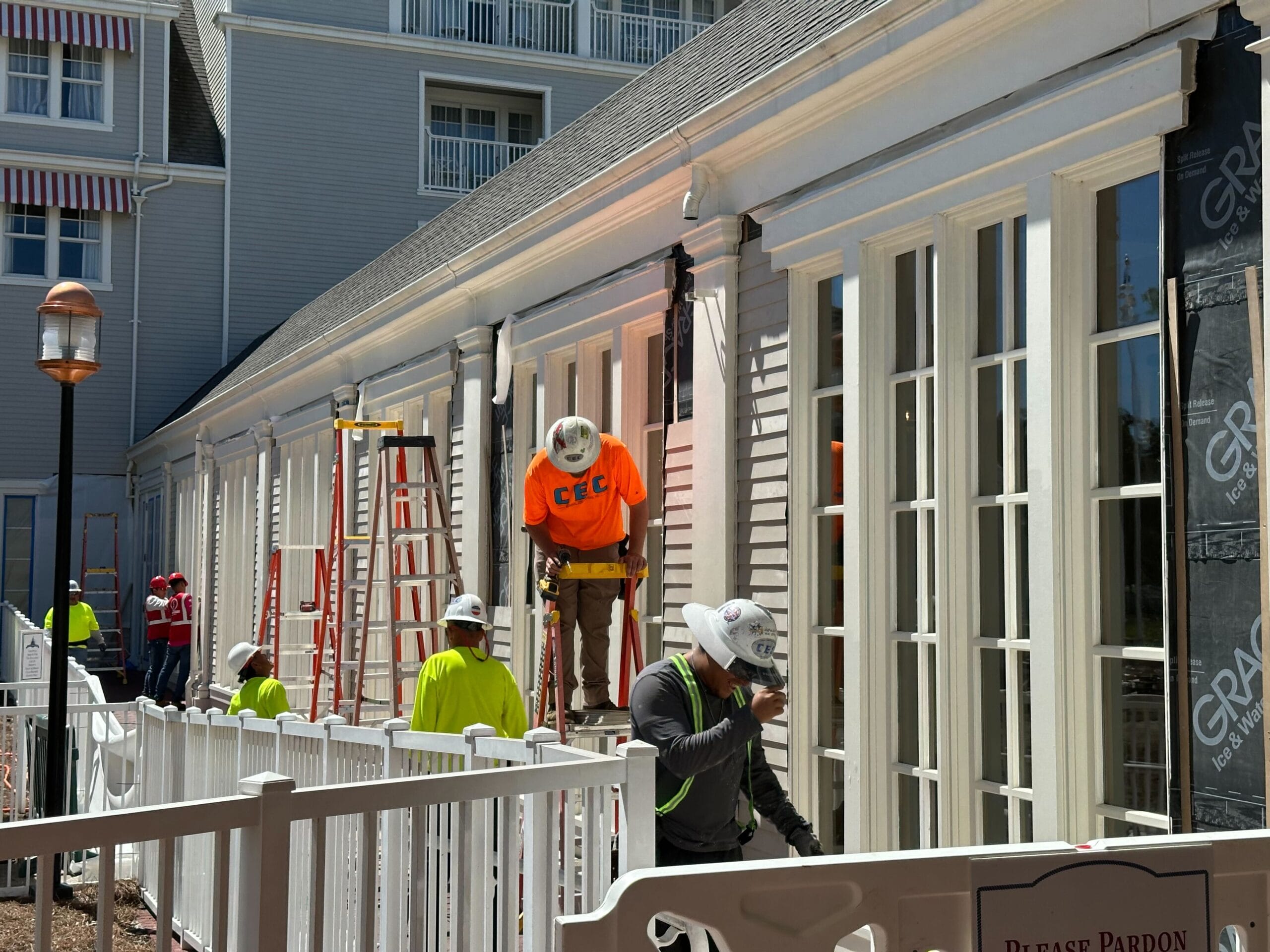 Workers in safety gear renovate a building's exterior with white siding at the edge of Stormalong Bay Pool. Ladders and scaffolding are in use, and a barrier ensures safety for those visiting Disney’s Yacht Club during construction.