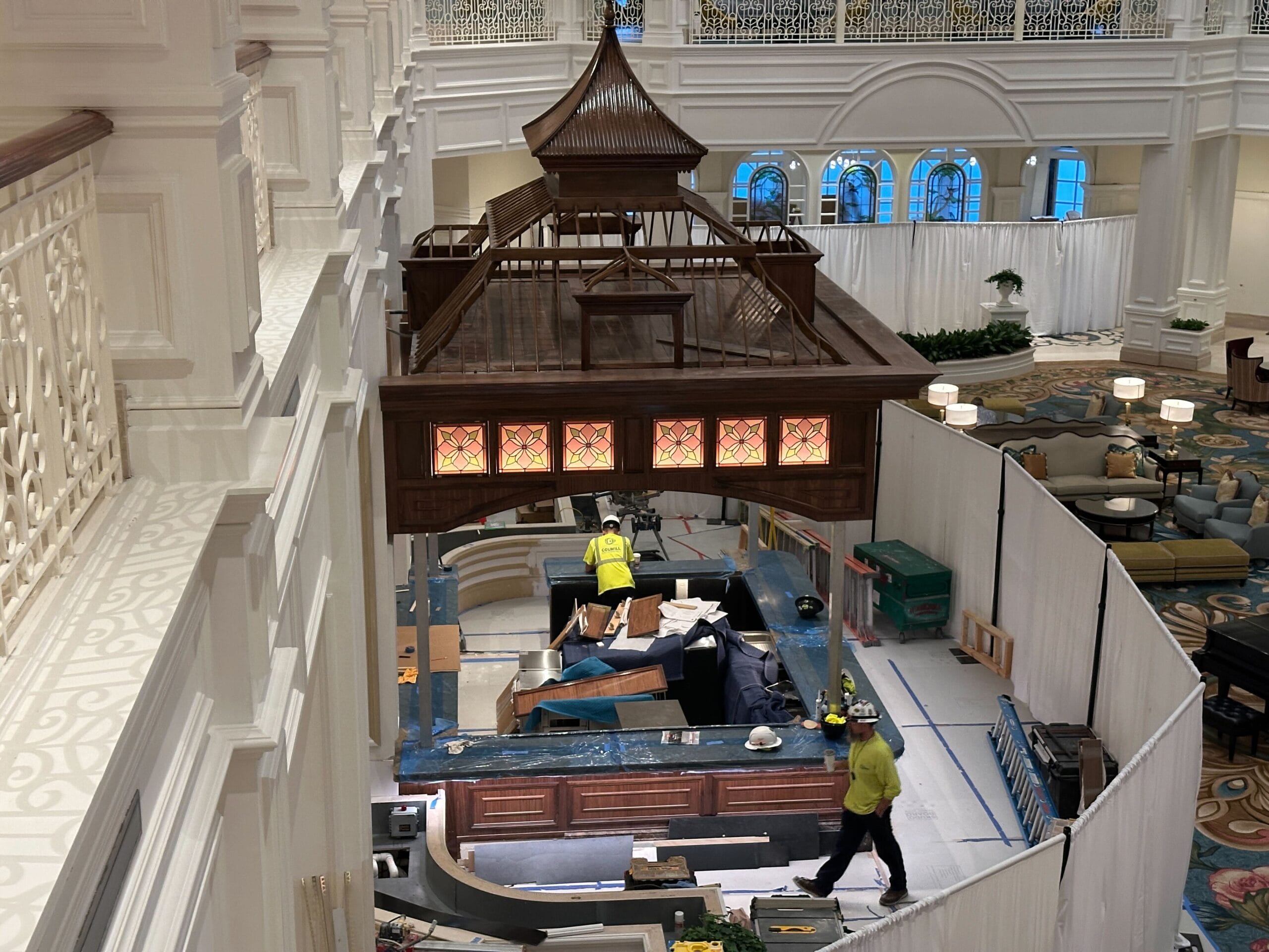 Two workers renovate a lobby with a wooden roof near Tea Room windows in a grand hall, similar to Disney Park resort elegance.
