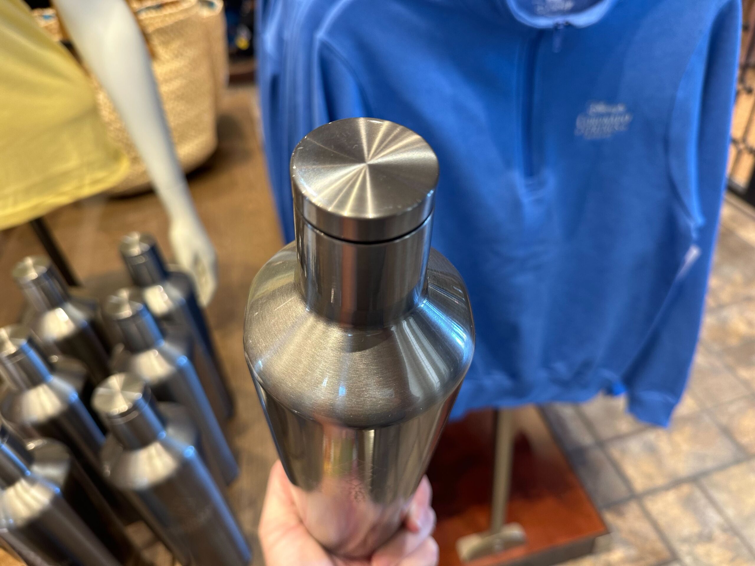 Close-up of a stainless steel water bottle branded with Coronado Springs Merchandise, held in a hand. In the background, a soft blue sweatshirt drapes casually alongside more bottles, evoking the perfect blend of style and practicality.