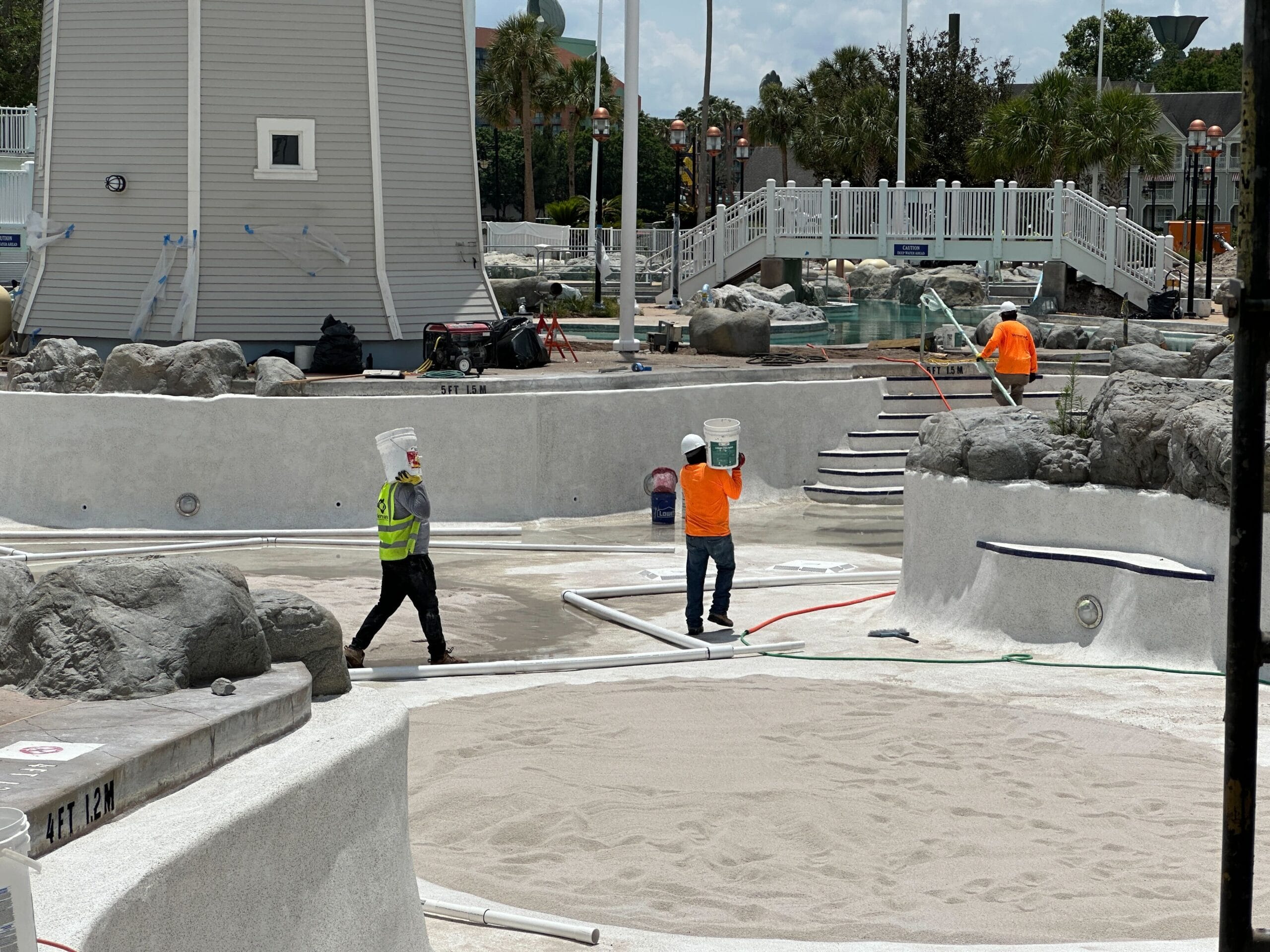 Workers in safety gear renovate an empty pool, carrying buckets and tools near artificial rocks and a lighthouse structure.
