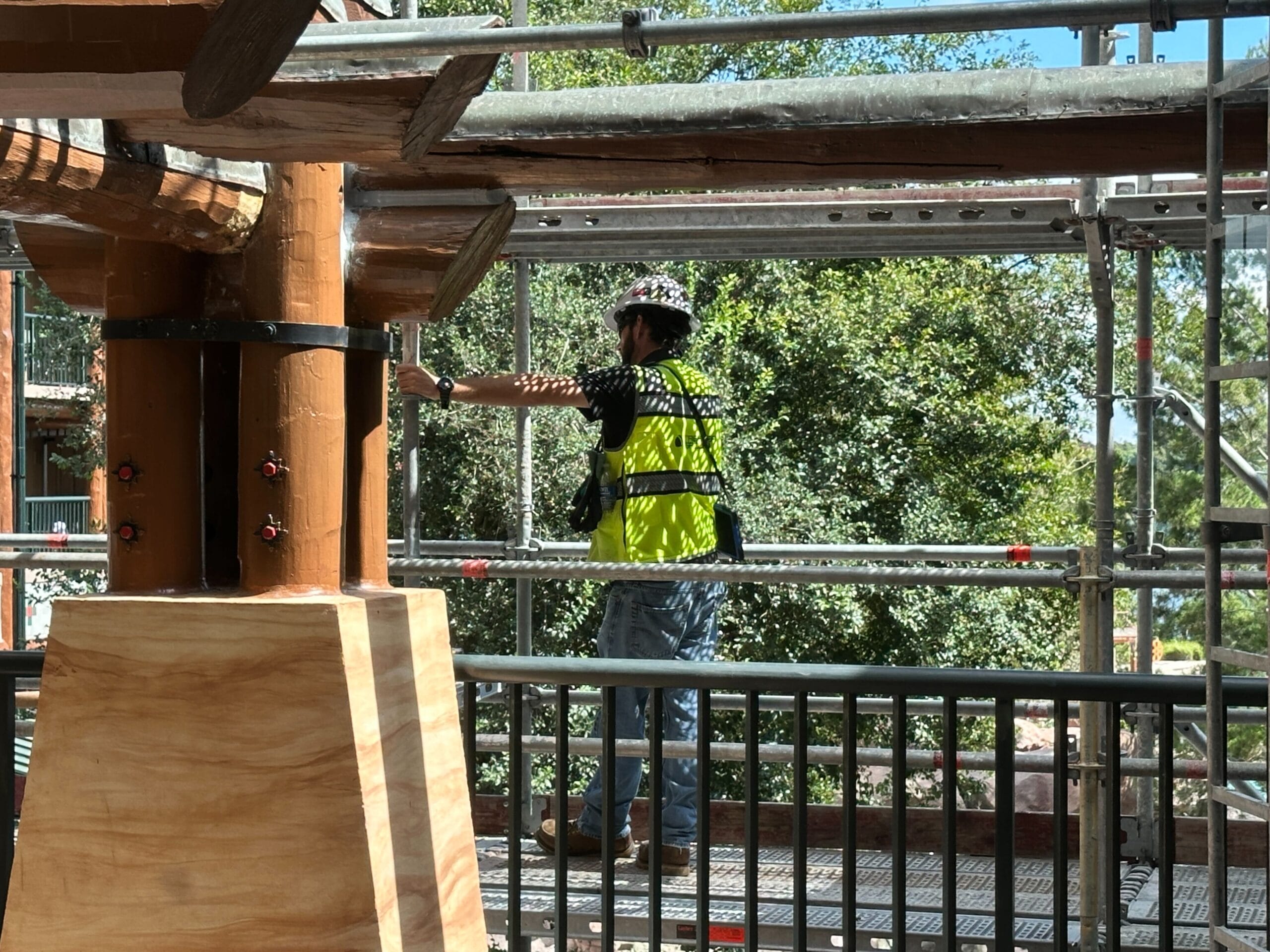 A construction worker in a yellow vest stands on scaffolding at Fort Wilderness Lodge near wooden beams and green trees.
