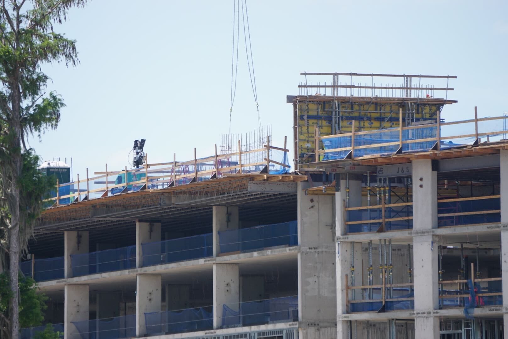 A multi-story building under construction, with scaffolding, blue netting, and exposed concrete beams. A crane hook and construction materials are visible on the top floor.