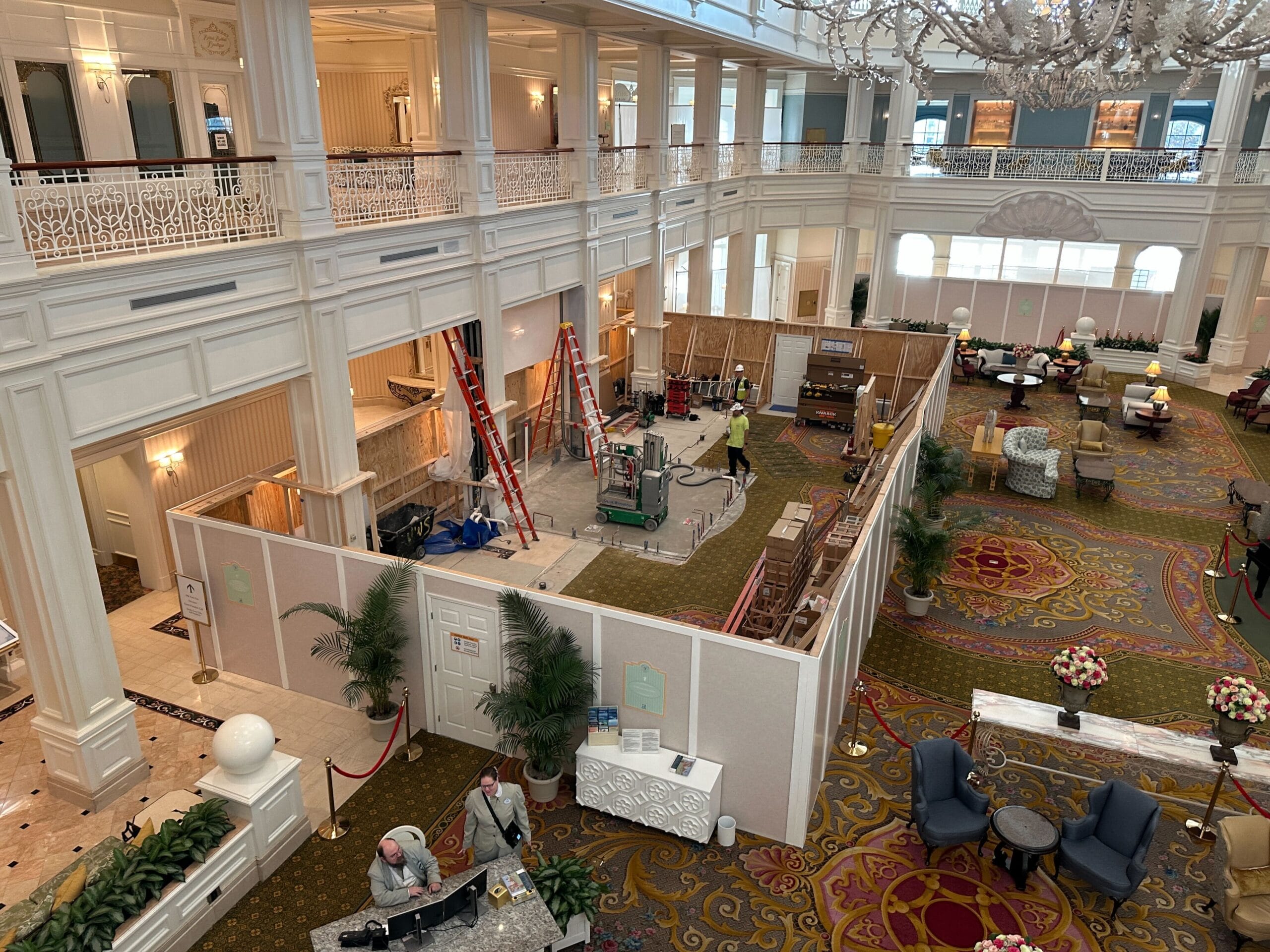 Large hotel lobby with construction barriers and equipment, workers present, and guests sitting in a lounge area nearby.