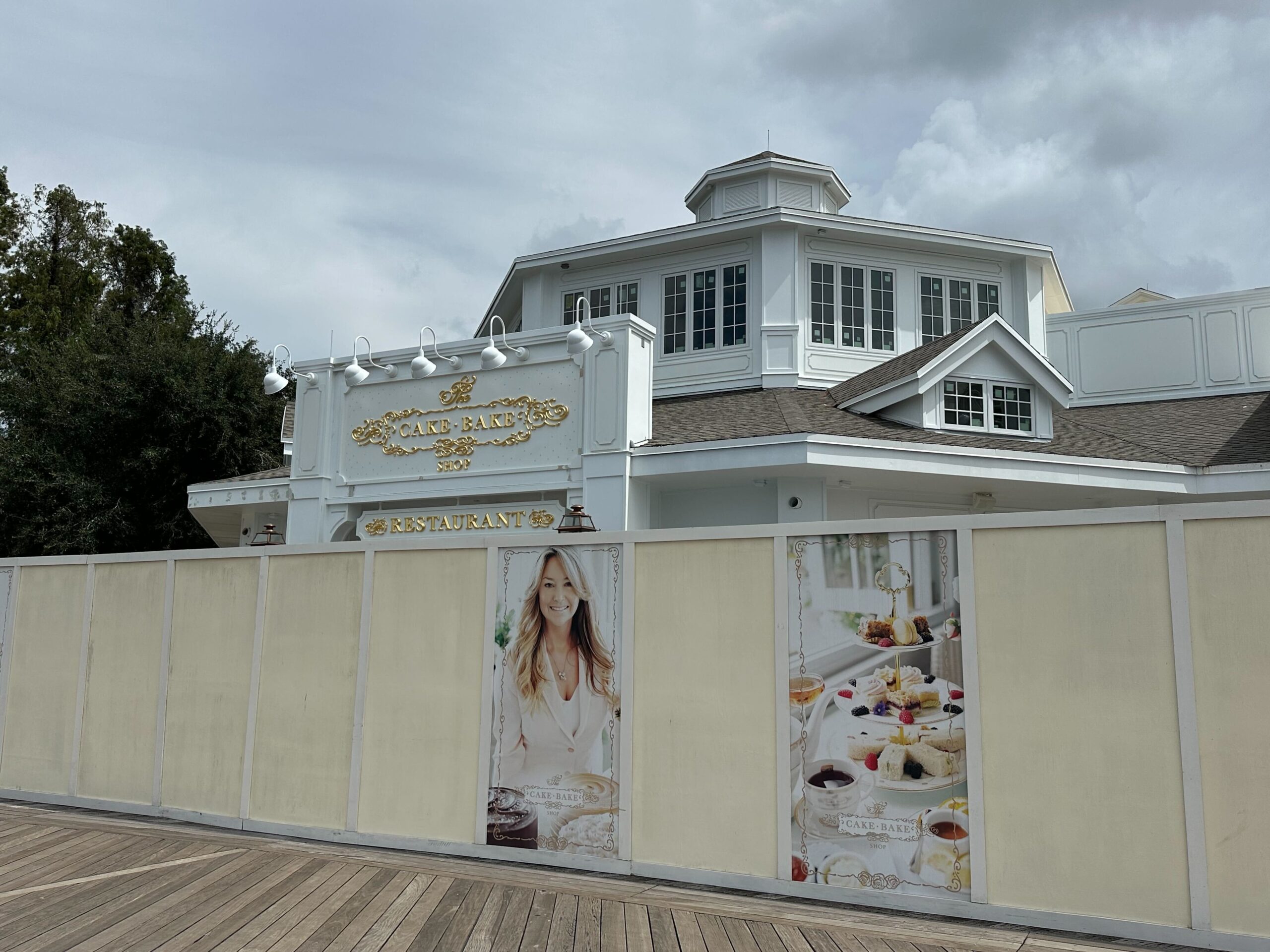 A white, two-story building with a sign that reads Cake Bake Shop is under construction, surrounded by a white fence with images of desserts and a smiling woman.