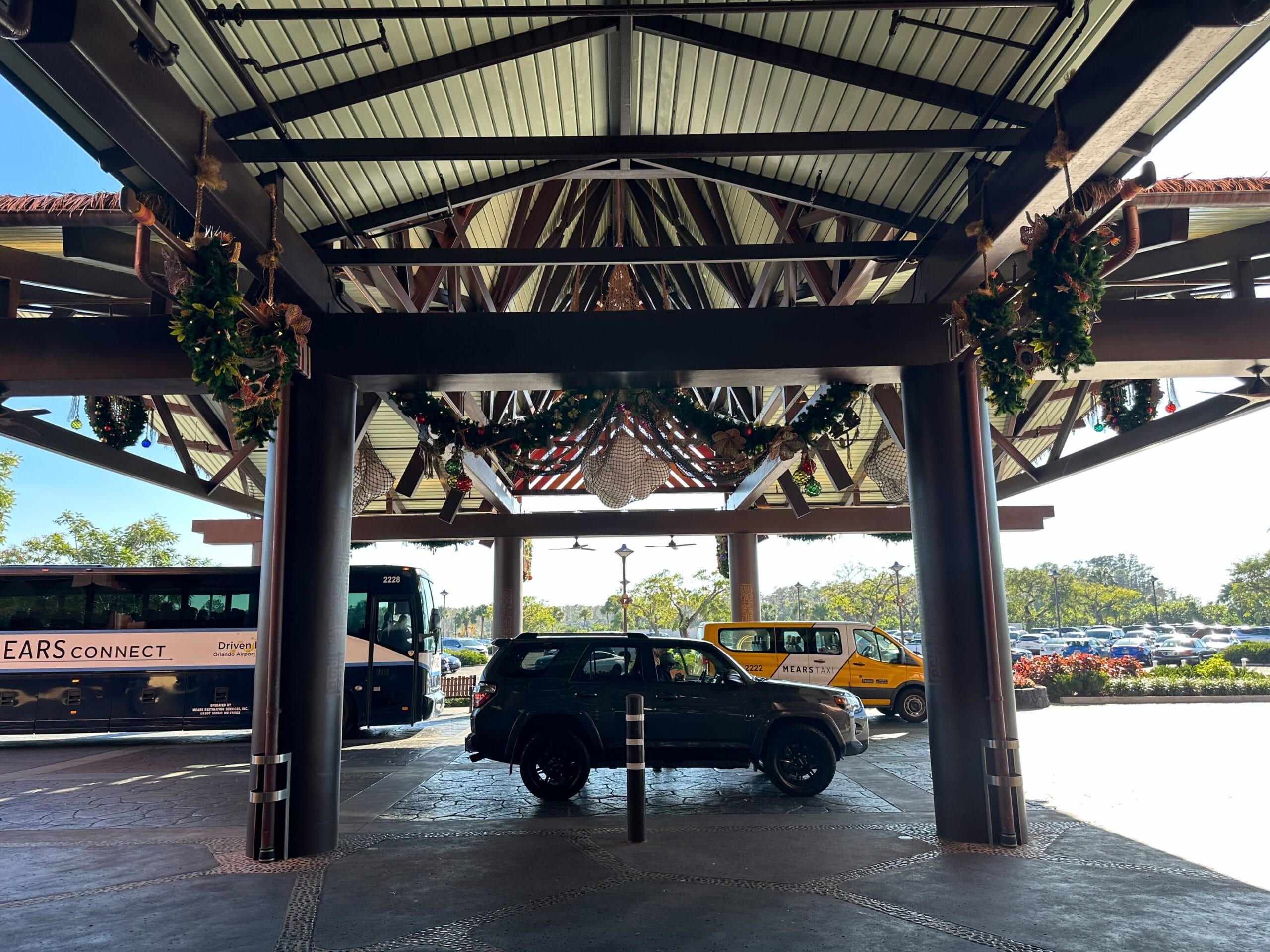 Covered area with a parked bus and SUV, and a yellow taxi in the background. Hanging decorations are visible on the ceiling.