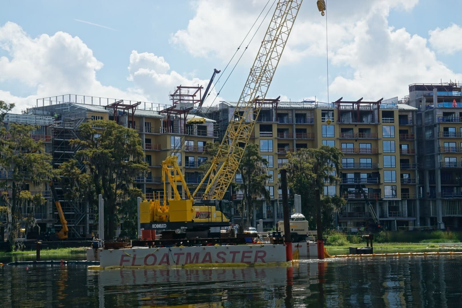 A yellow crane on a barge labeled FLOATMASTER operates near Disney Lakeshore Lodge by the water.