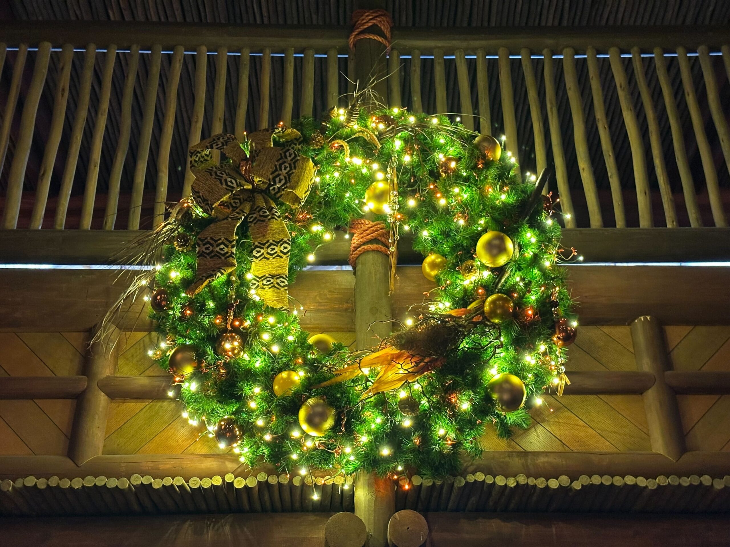 A large Christmas wreath with lights, gold ornaments, and a patterned bow hanging on a wooden wall.