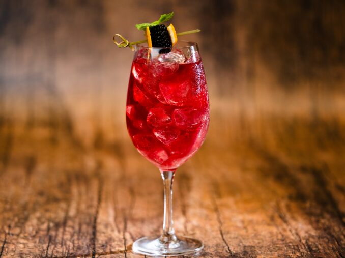 A tall glass filled with a red cocktail, garnished with a lemon twist, mint leaf, and a small black sphere, sits on a wooden surface—reminiscent of the whimsical concoctions enjoyed at Walt Disney World.