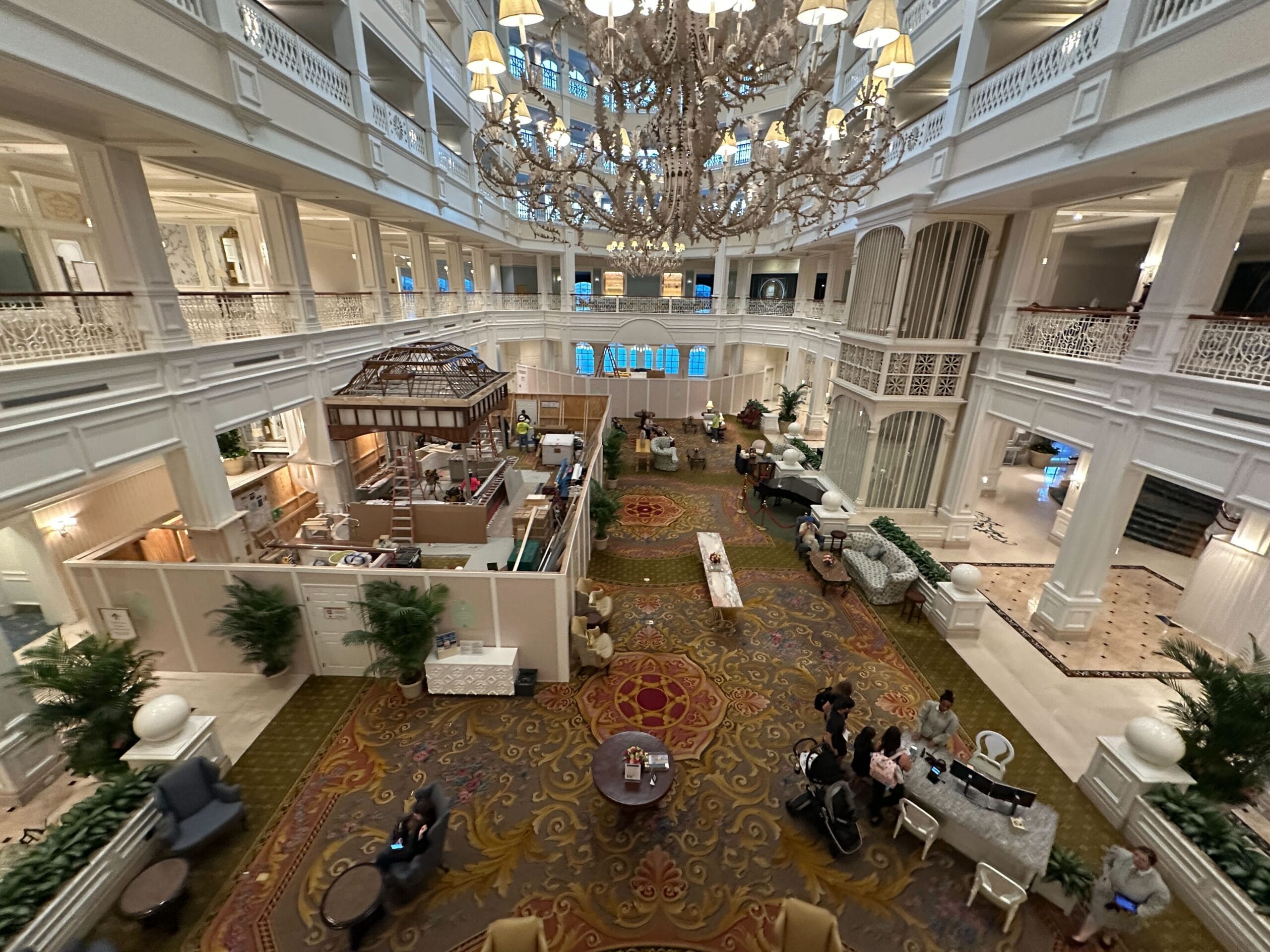 Elegant Grand Floridian hotel lobby with grand chandelier, ornate railings, plush seating, plants, and guests gathered below.