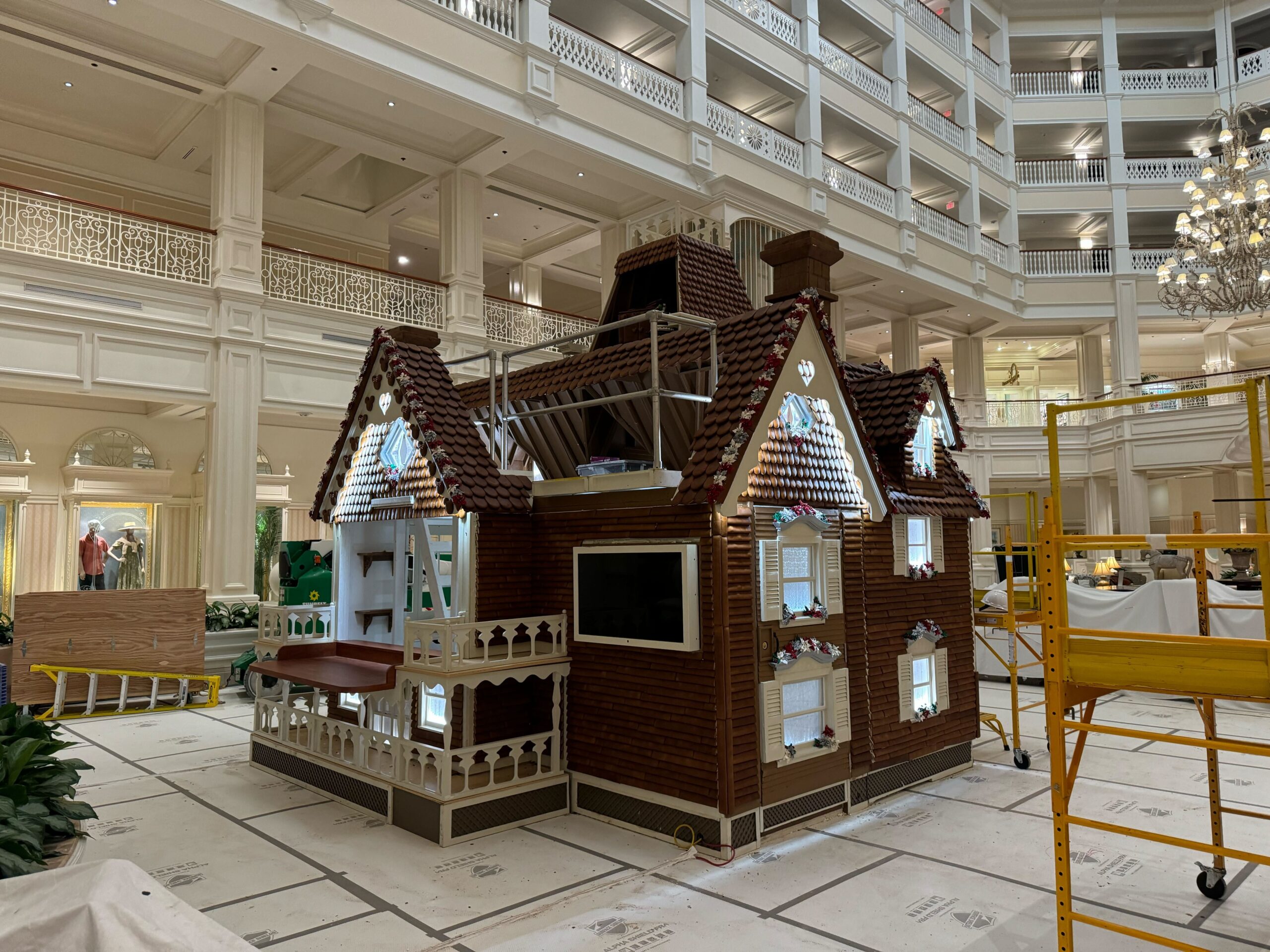 A large gingerbread house display is under construction in the grand lobby of Disney's Grand Floridian Resort, adorned with elegant railings and chandeliers, surrounded by scaffolding and construction materials.
