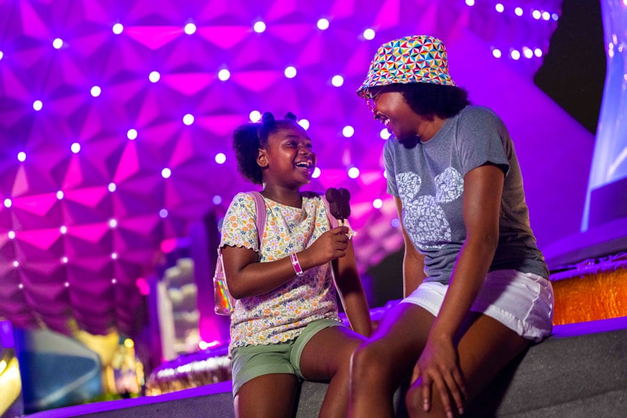 Family enjoying unlimited snacks, including Mickey ice cream bar sitting in front of Spaceship Earth at night during Disney After Hours Event in EPCOT