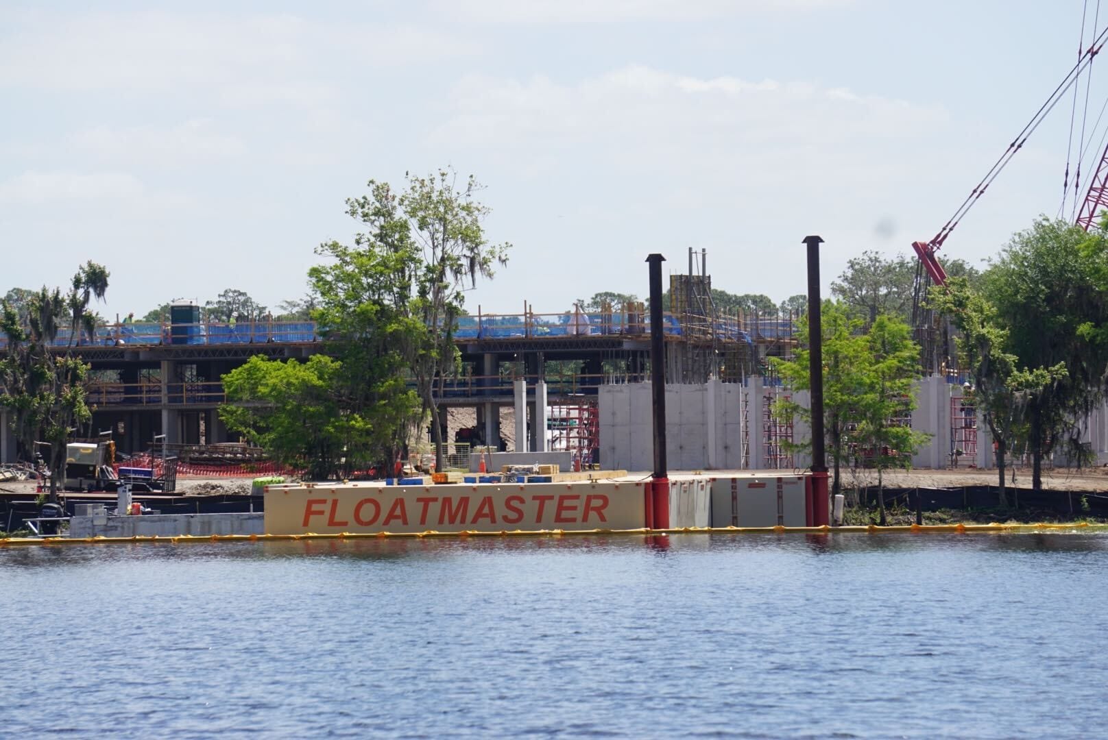 Construction site by a lake with a large barge labeled FLOATMASTER in the foreground. Building structures and cranes are visible in the background.