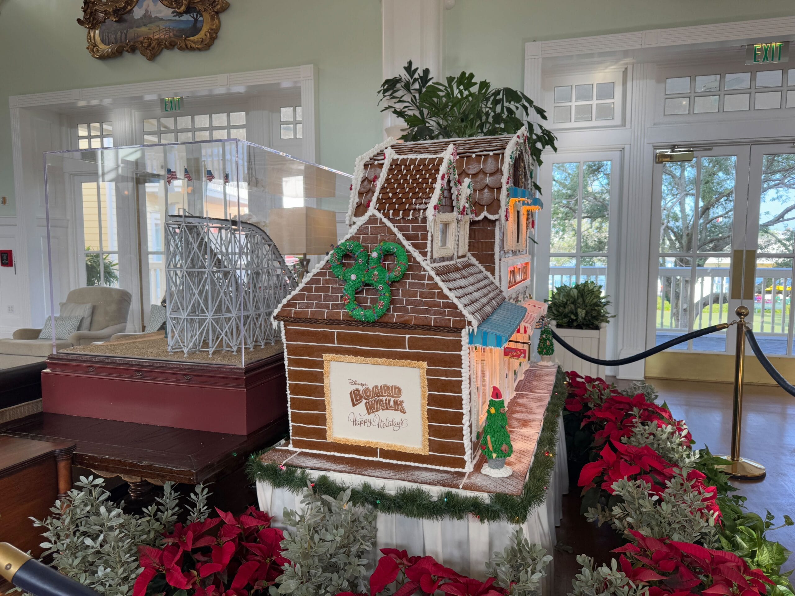 A festive gingerbread house display with a small roller coaster model beside it. Surrounded by red poinsettias and guarded by ropes. A windowed room with light streaming in.