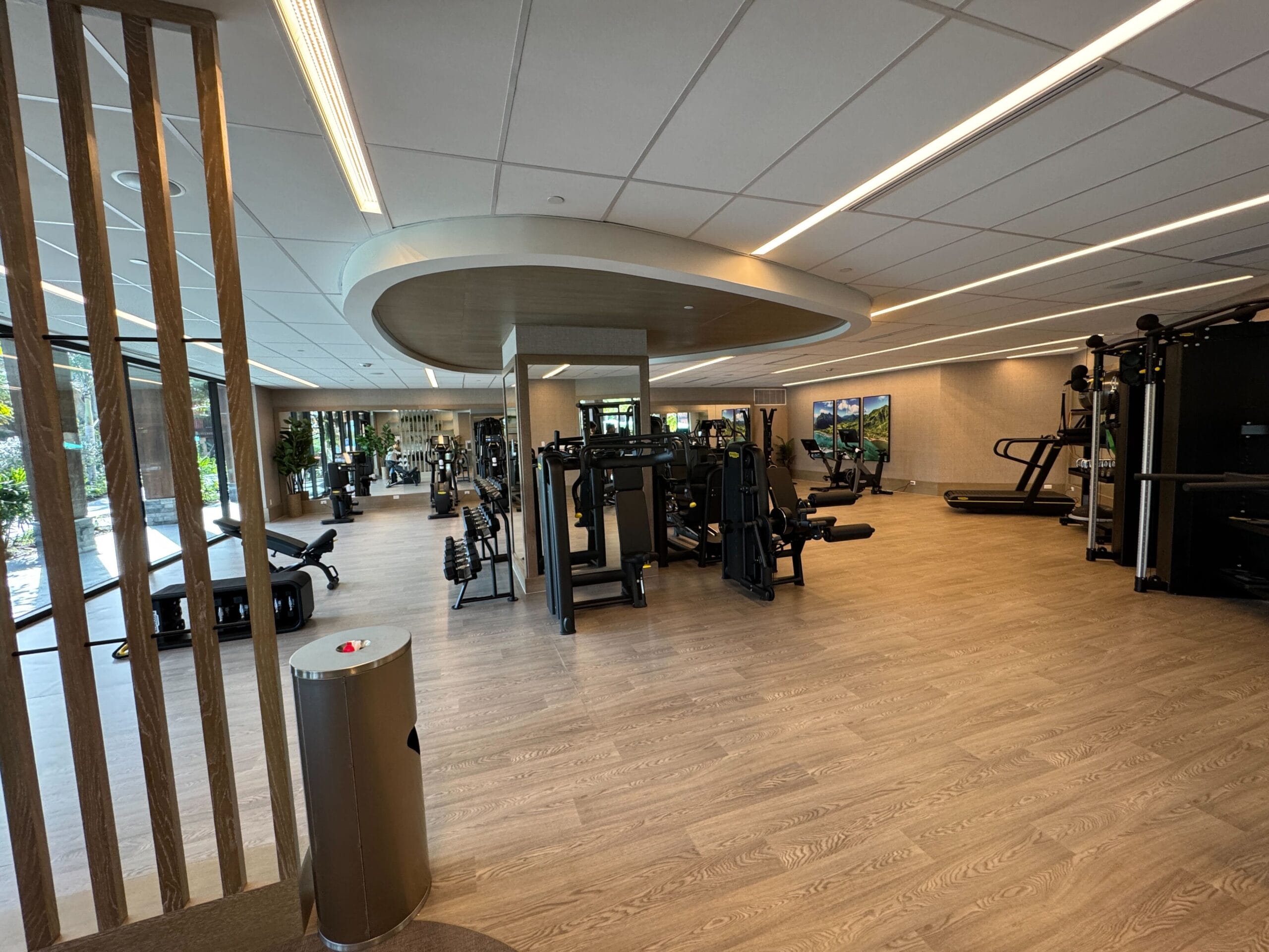 A modern gym with exercise machines, free weights, and treadmills. Large windows line one wall, and natural light illuminates the wooden floor.