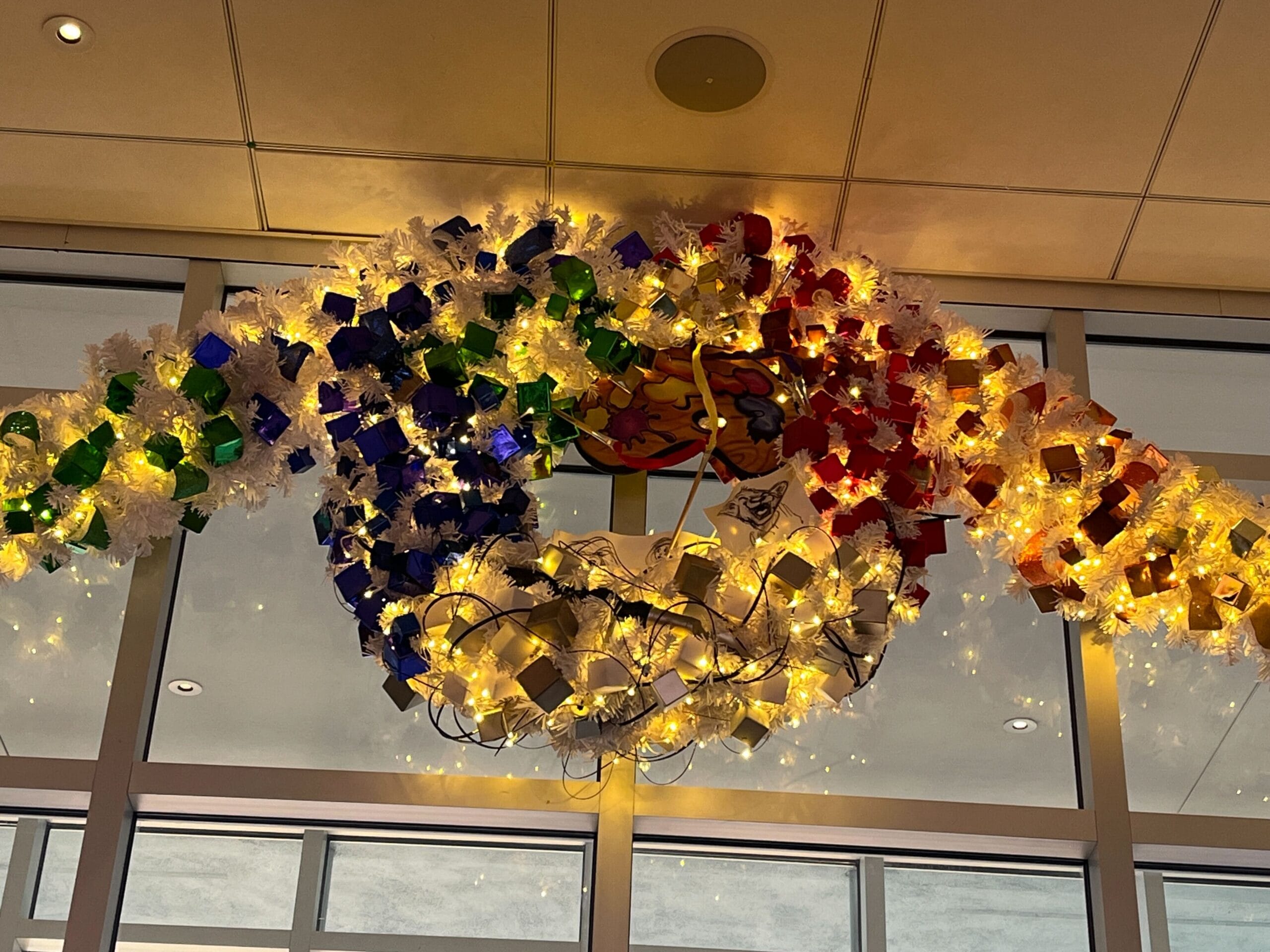 A circular ceiling decoration with colorful flowers and lights is hanging against a window.