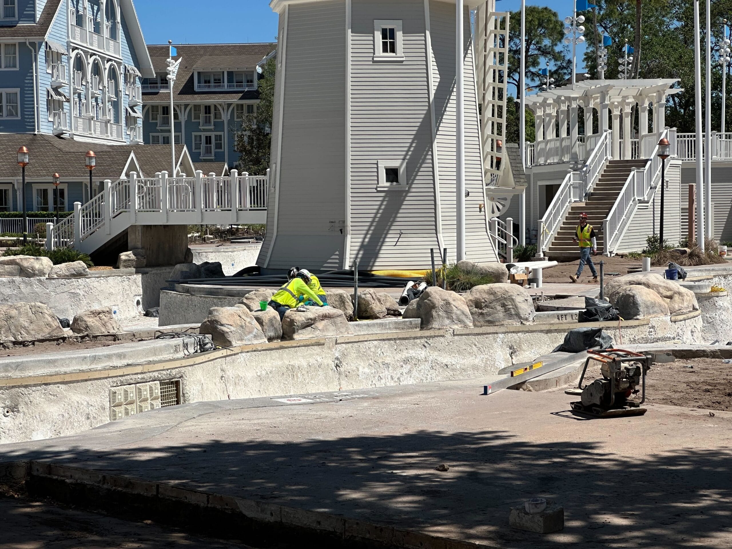 Construction workers in bright clothing are busy renovating the drained Stormalong Bay Pool near Disney’s Yacht Club, surrounded by a white wooden structure and multi-story buildings.