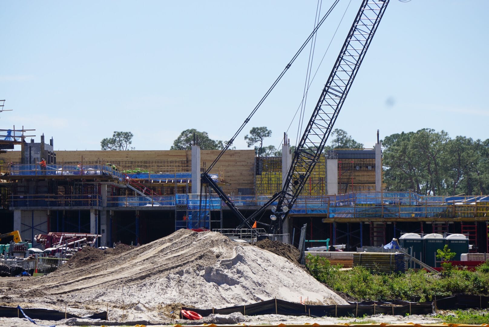 Construction site with a large crane, sand piles, and partially built structures in the background, surrounded by trees and under a clear sky.