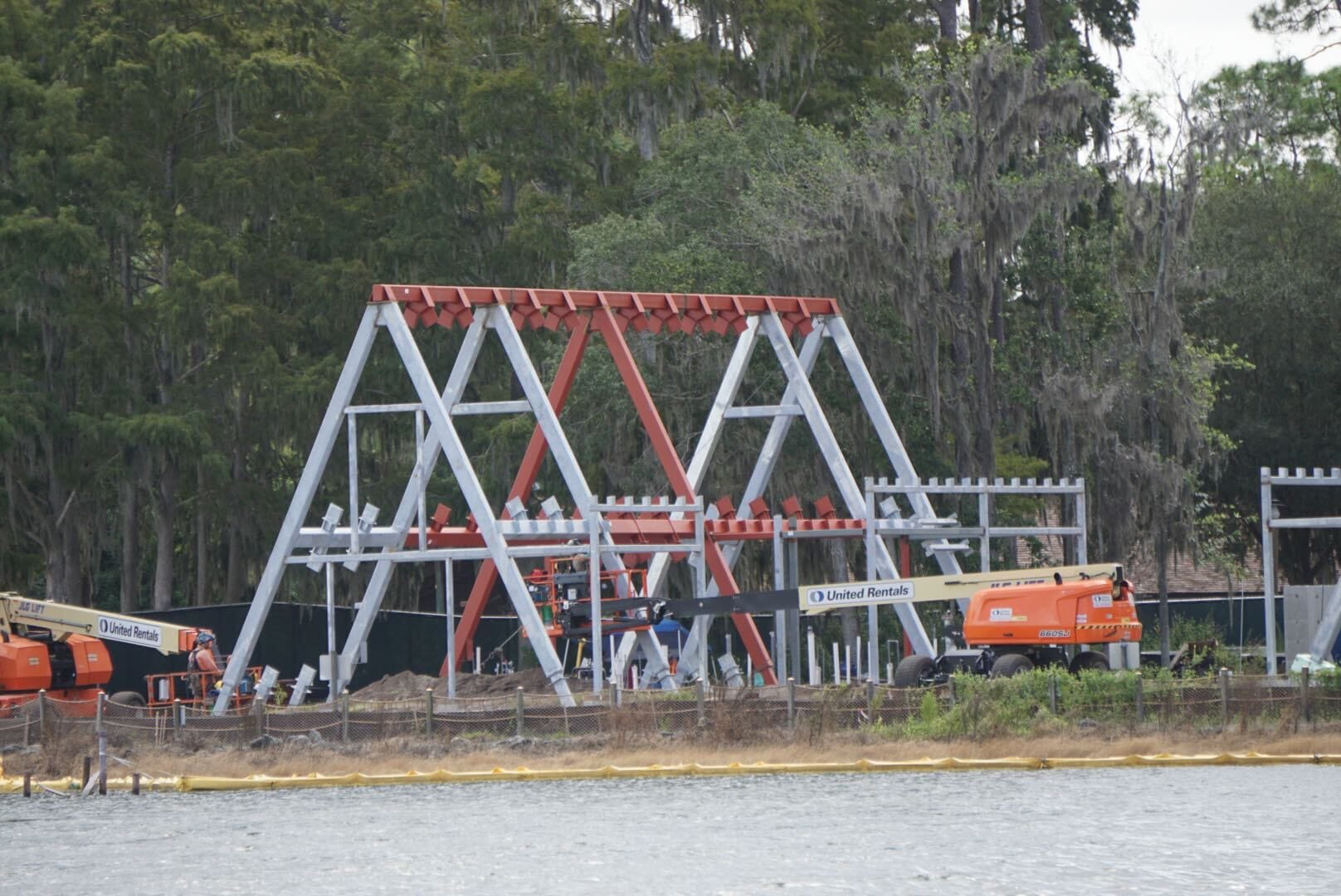 A red and white steel structure under construction near water at Lakeshore Lodge, surrounded by trees and equipment.