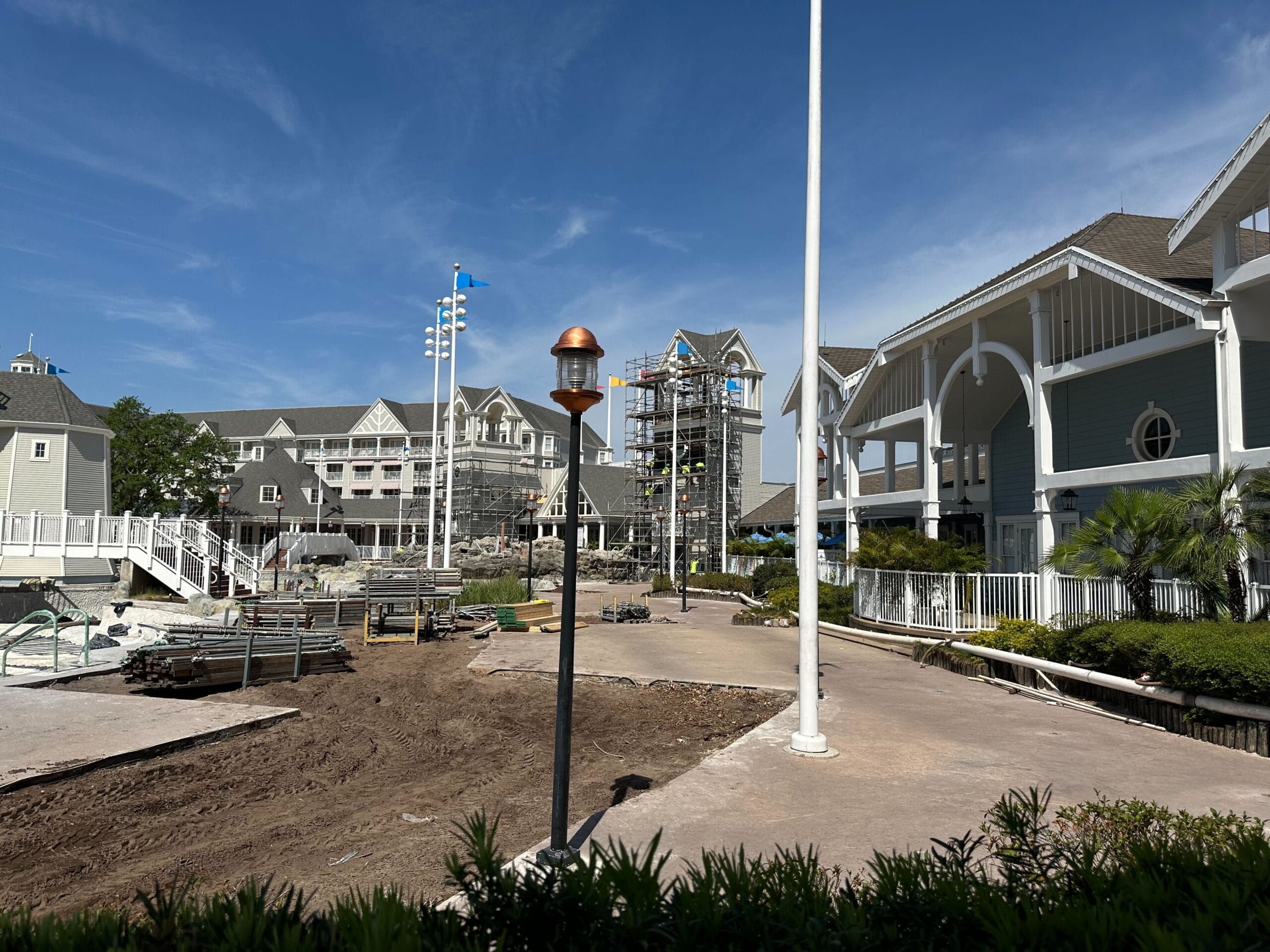 A construction site buzzes with activity near a large white and gray building at Disney’s Yacht Club, featuring multiple sections, scaffolding, and equipment. The scene is framed by a blue sky and a lamppost in the foreground.