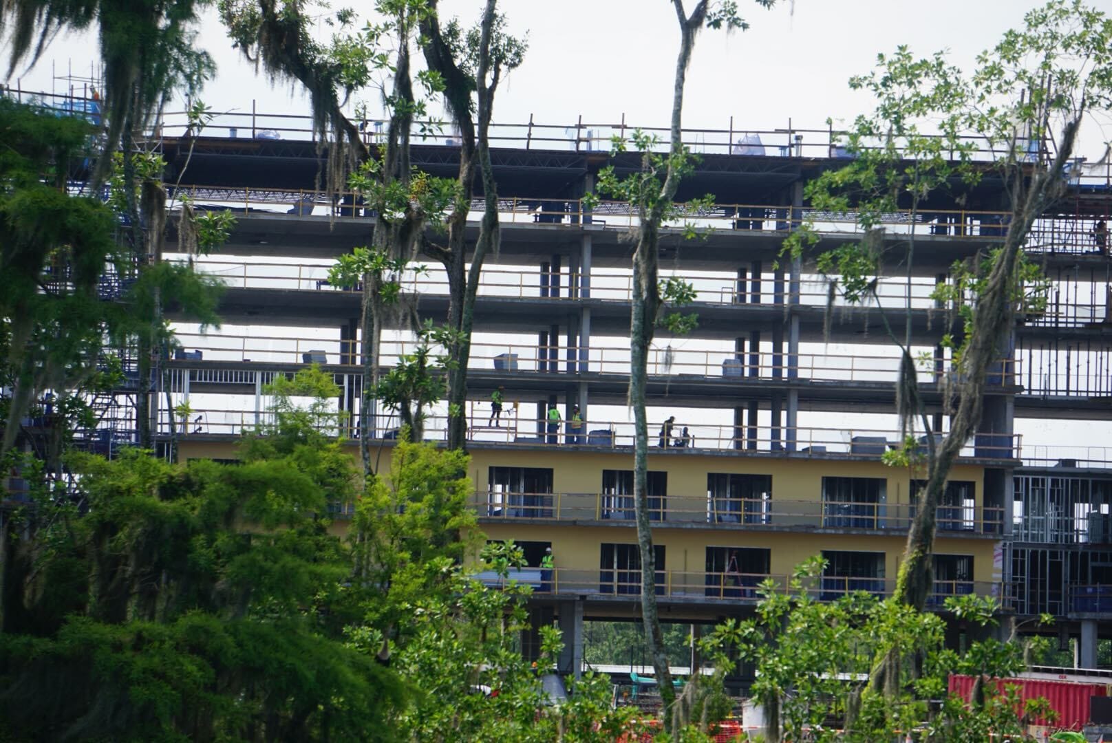 Multi-story building under construction, framed by green trees in the foreground.