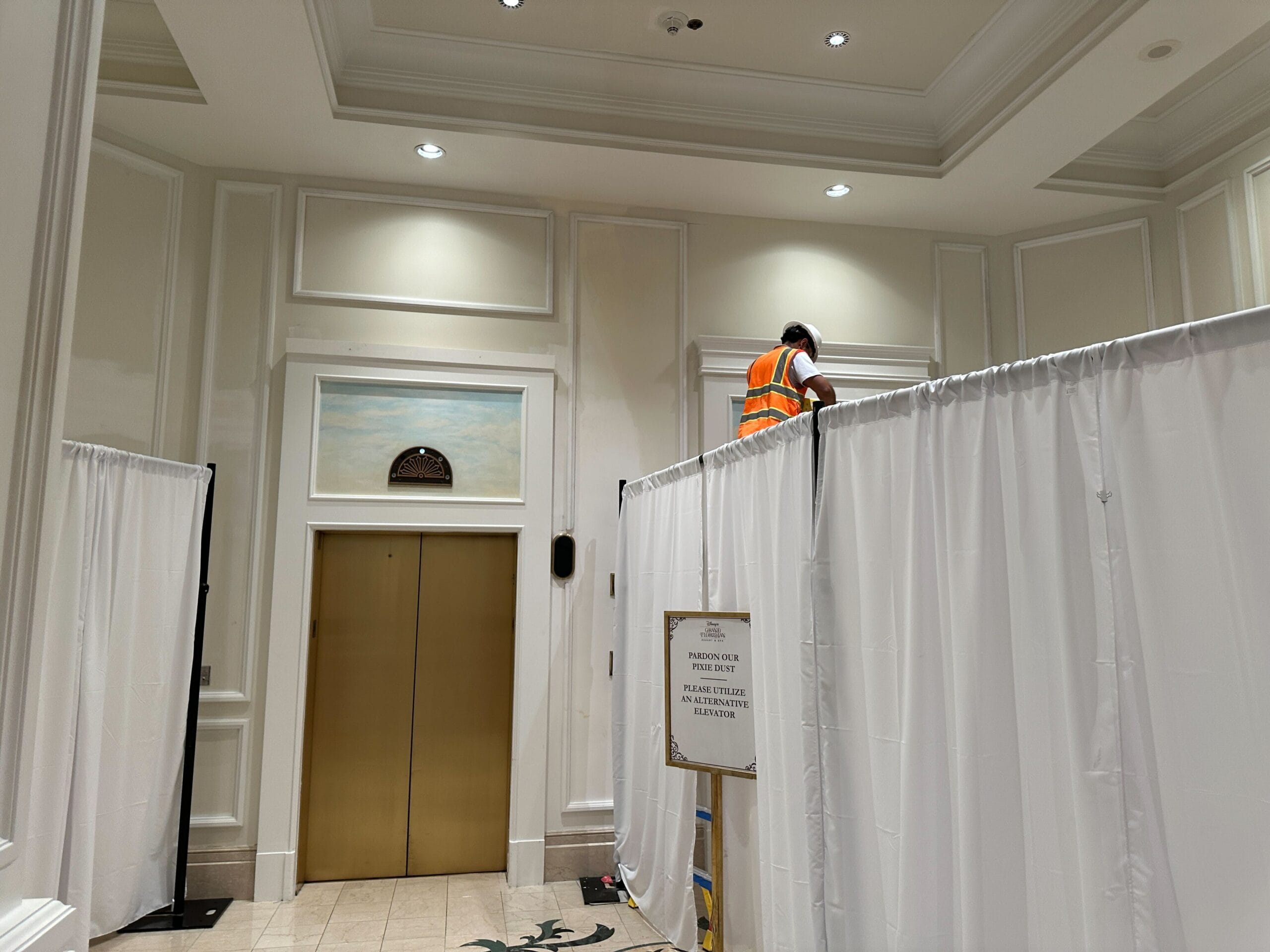 A worker in an orange vest stands on a ladder behind white curtains near an elevator in a building lobby.