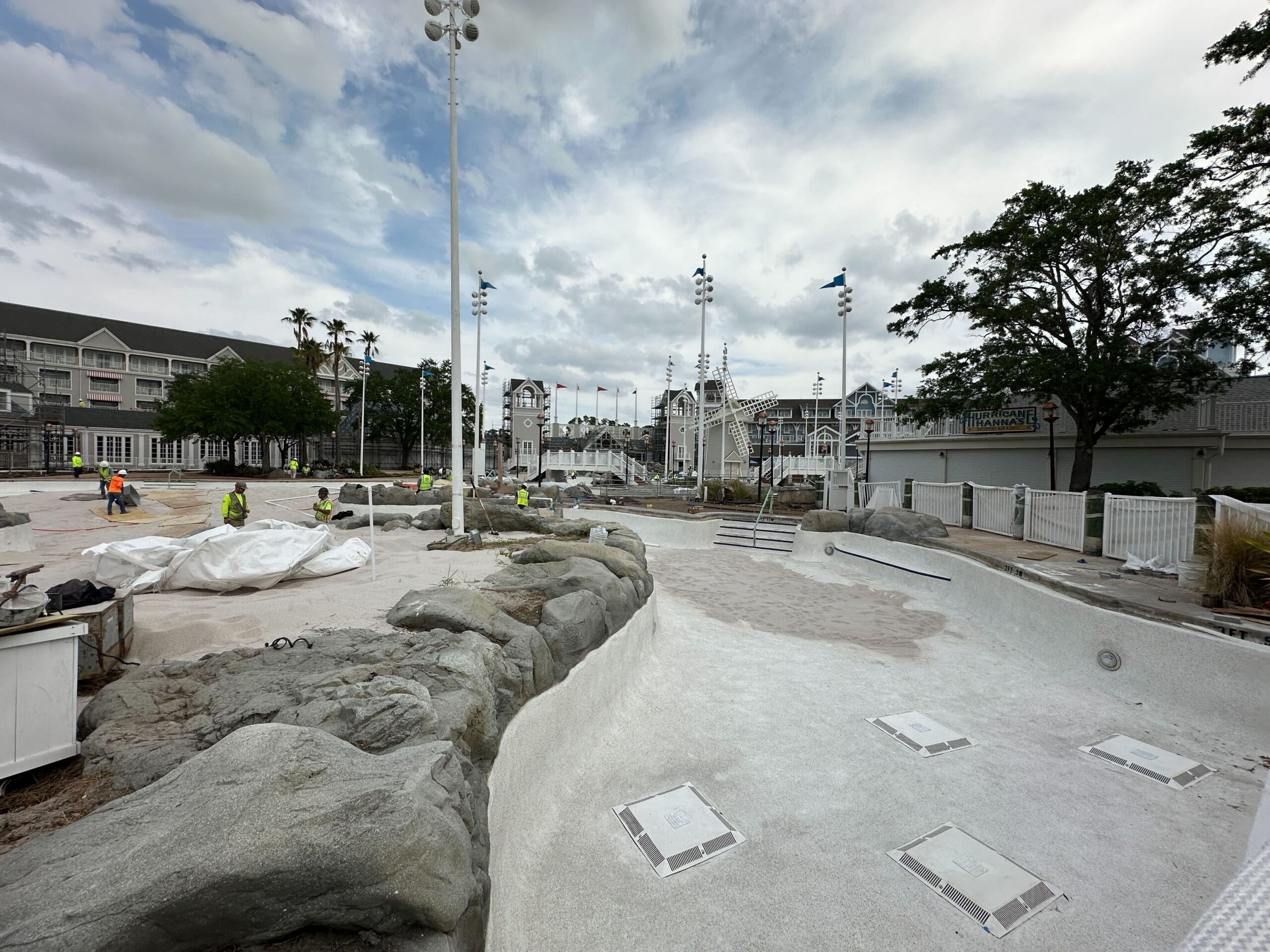 Empty swimming pool under construction with workers, rocks, and buildings in the background on a cloudy day.