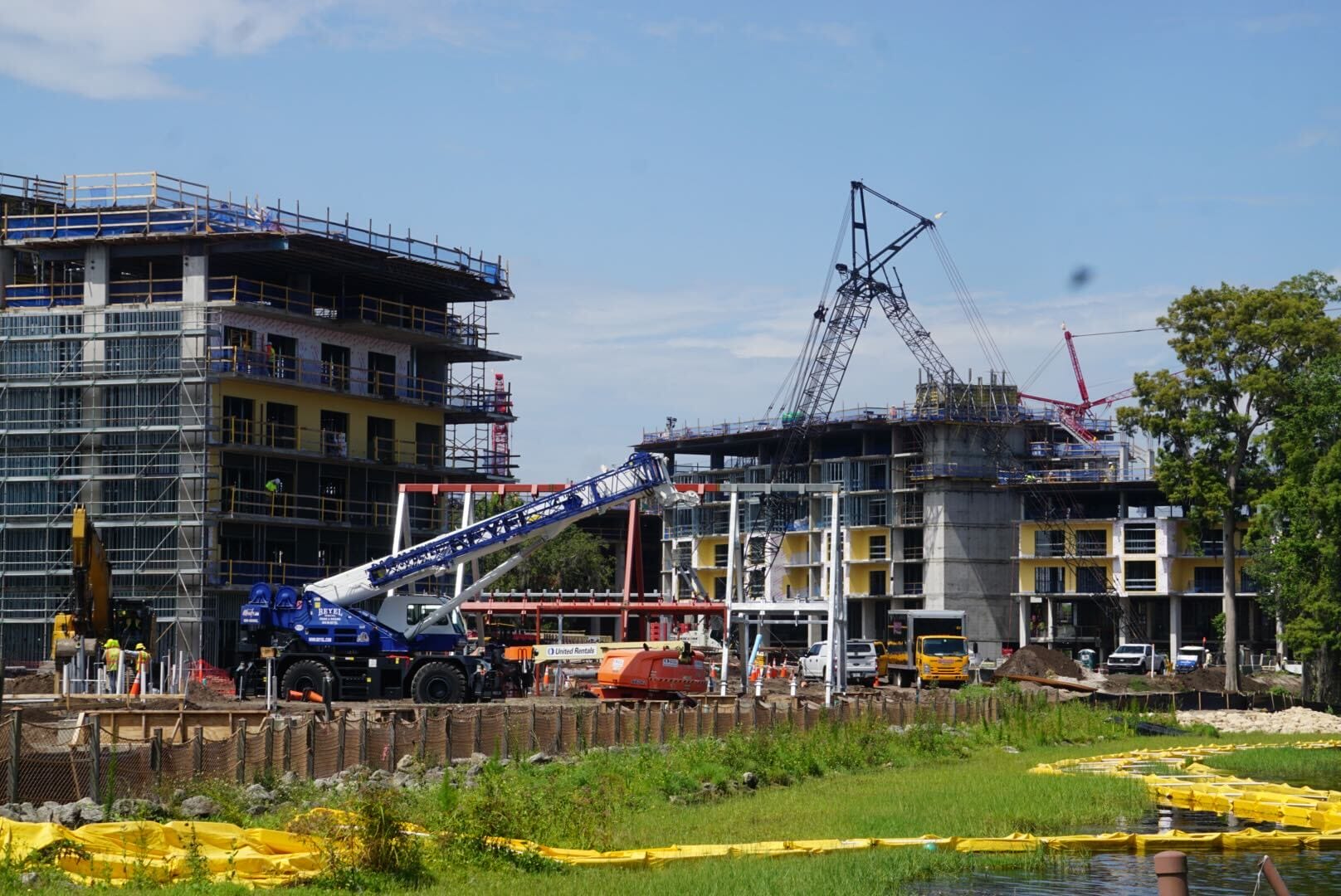 Construction site with cranes, scaffolding, and workers near a pond with yellow barriers and green grass in the foreground.