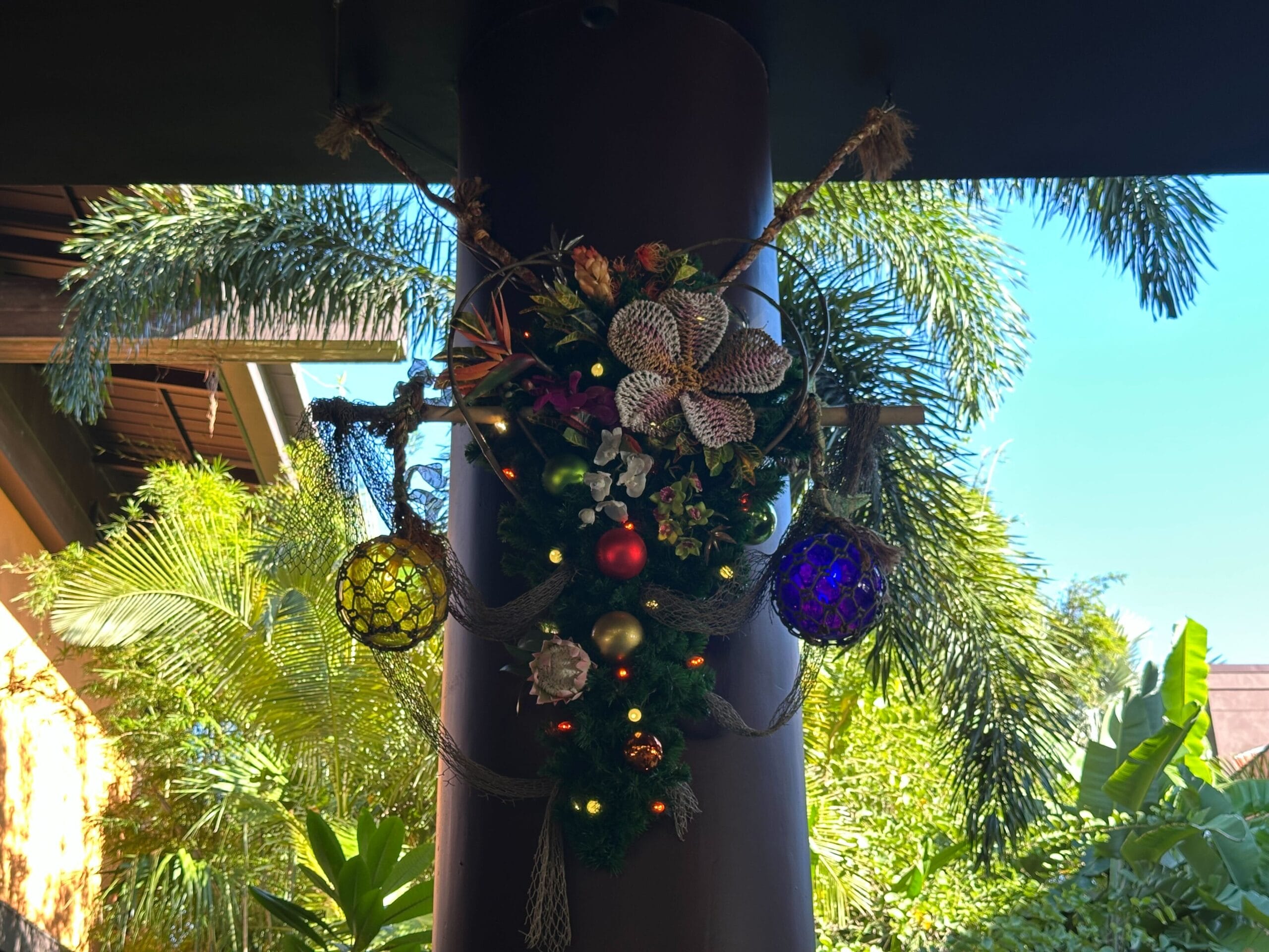 A decorative arrangement on a column features pine branches, ornaments, and lights. Tropical plants and a clear sky are in the background.