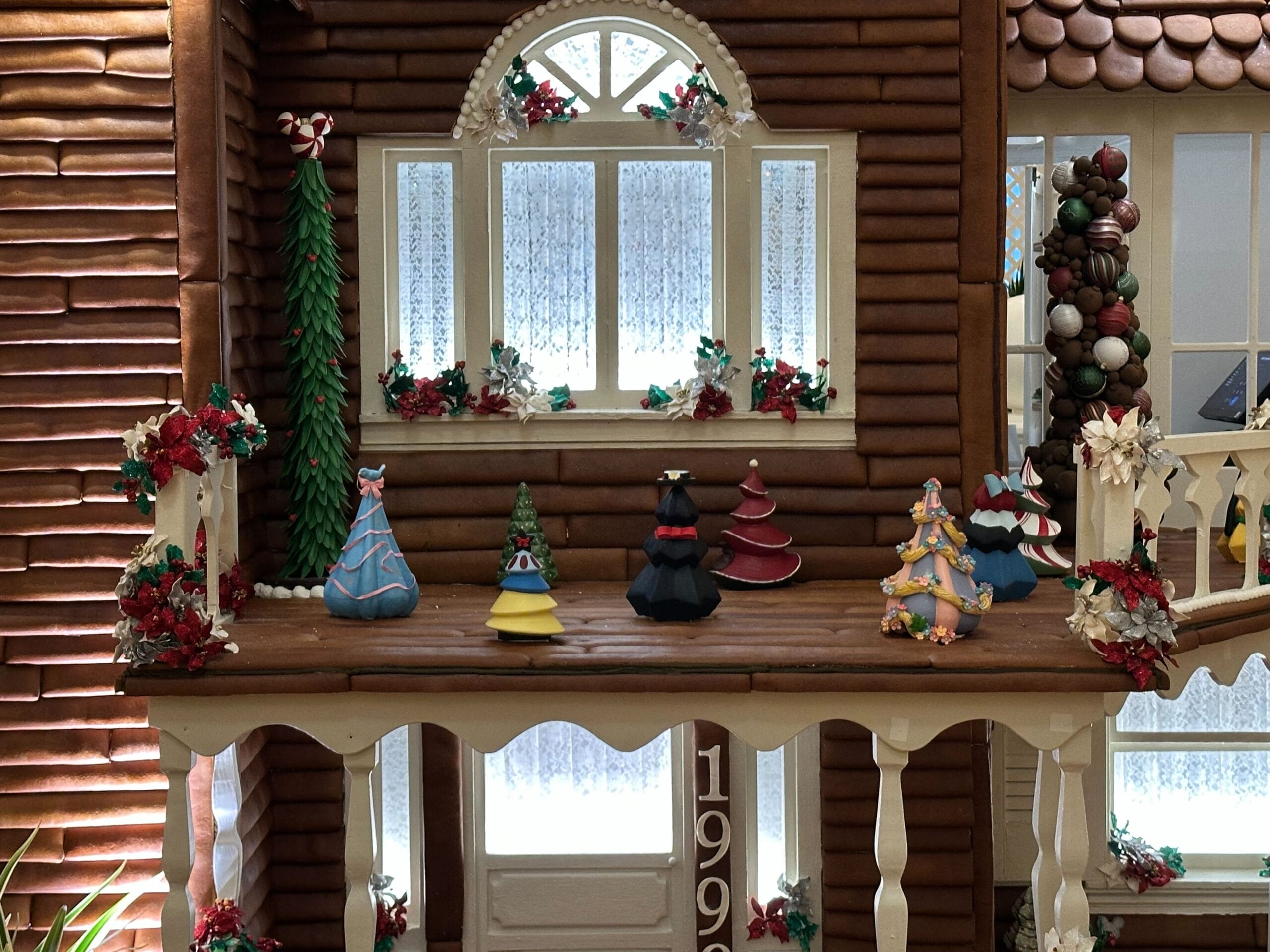 A gingerbread house display with colorful miniature Christmas trees and ornaments on a wooden porch.