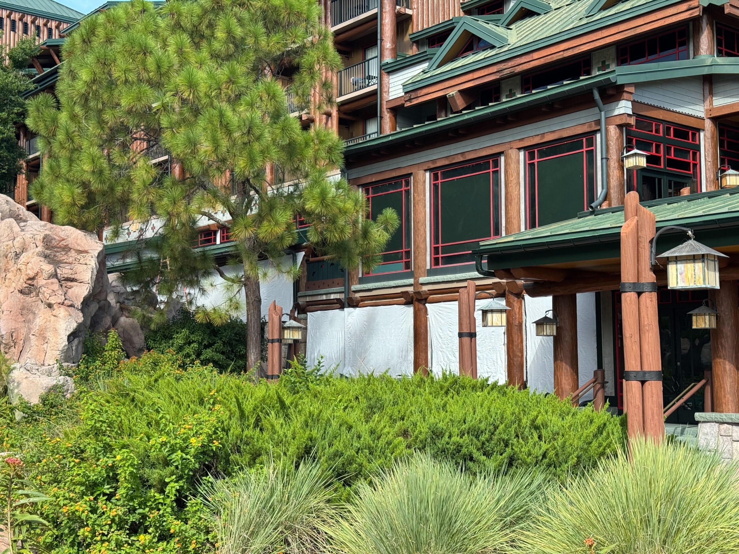 Lodge-style building with green roof, red accents, pine tree, and lush landscaping in front.
