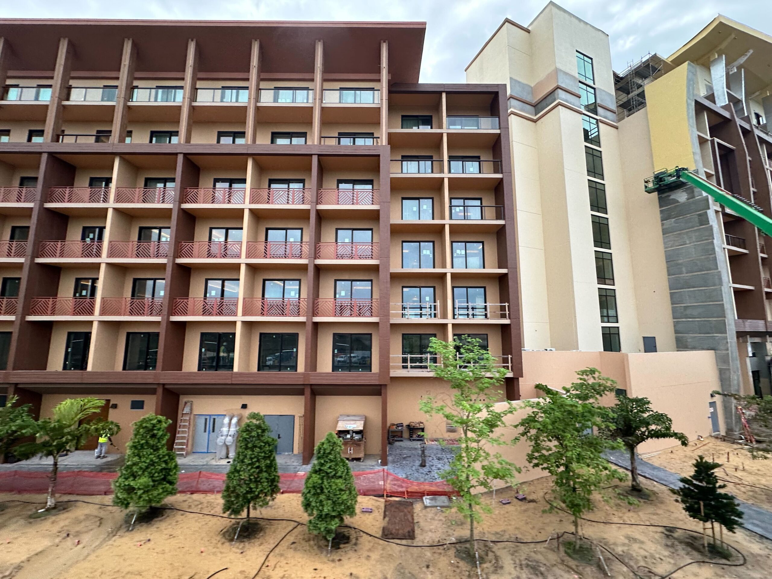 A view of a mid-rise building with multiple balconies and a construction site with a crane on the right side. The ground in front has newly planted trees and visible red construction barriers.