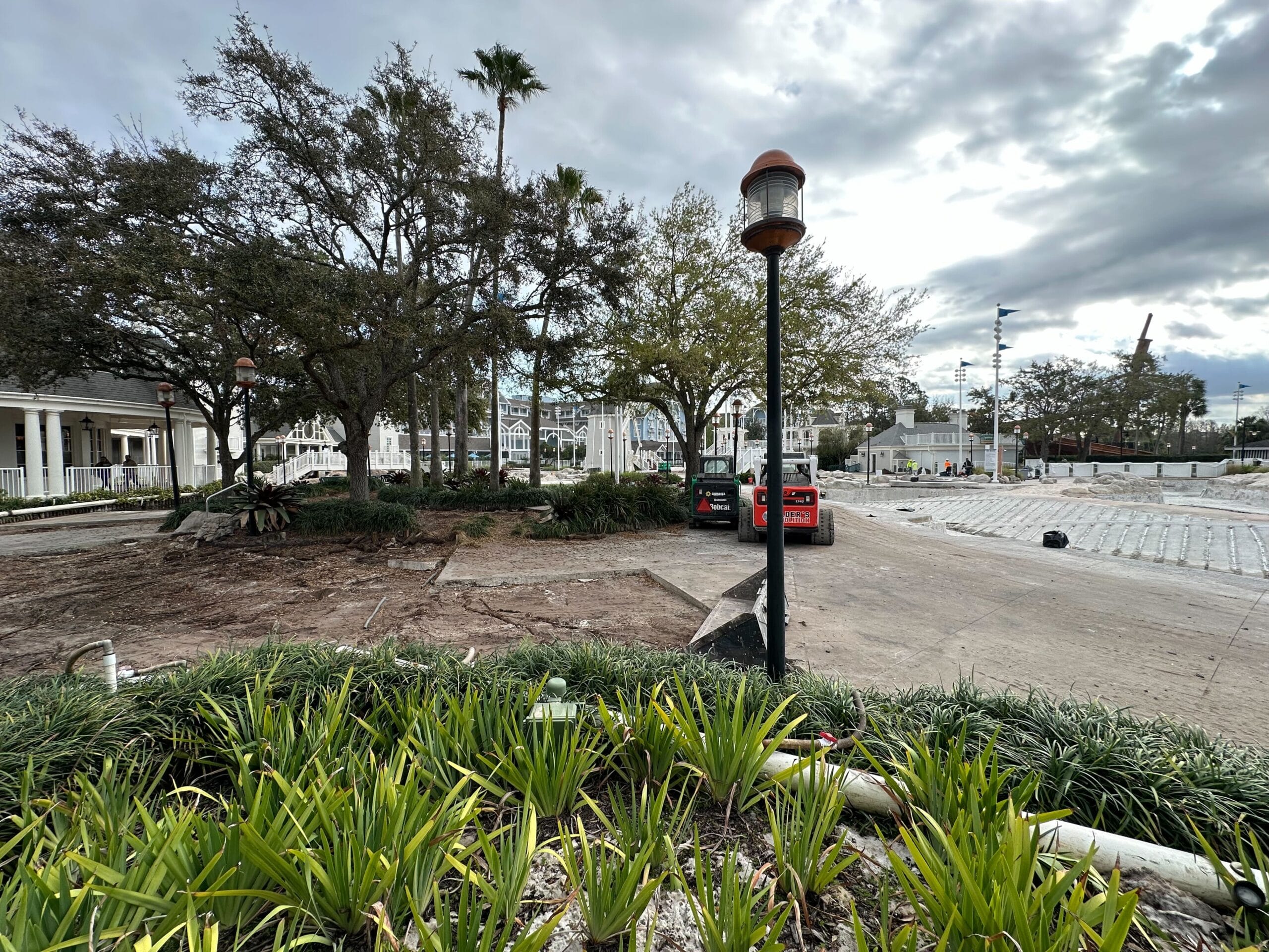 Construction scene with a partially paved area, several trees, two parked cars, and a lamp post. Overcast sky and some plants in the foreground.