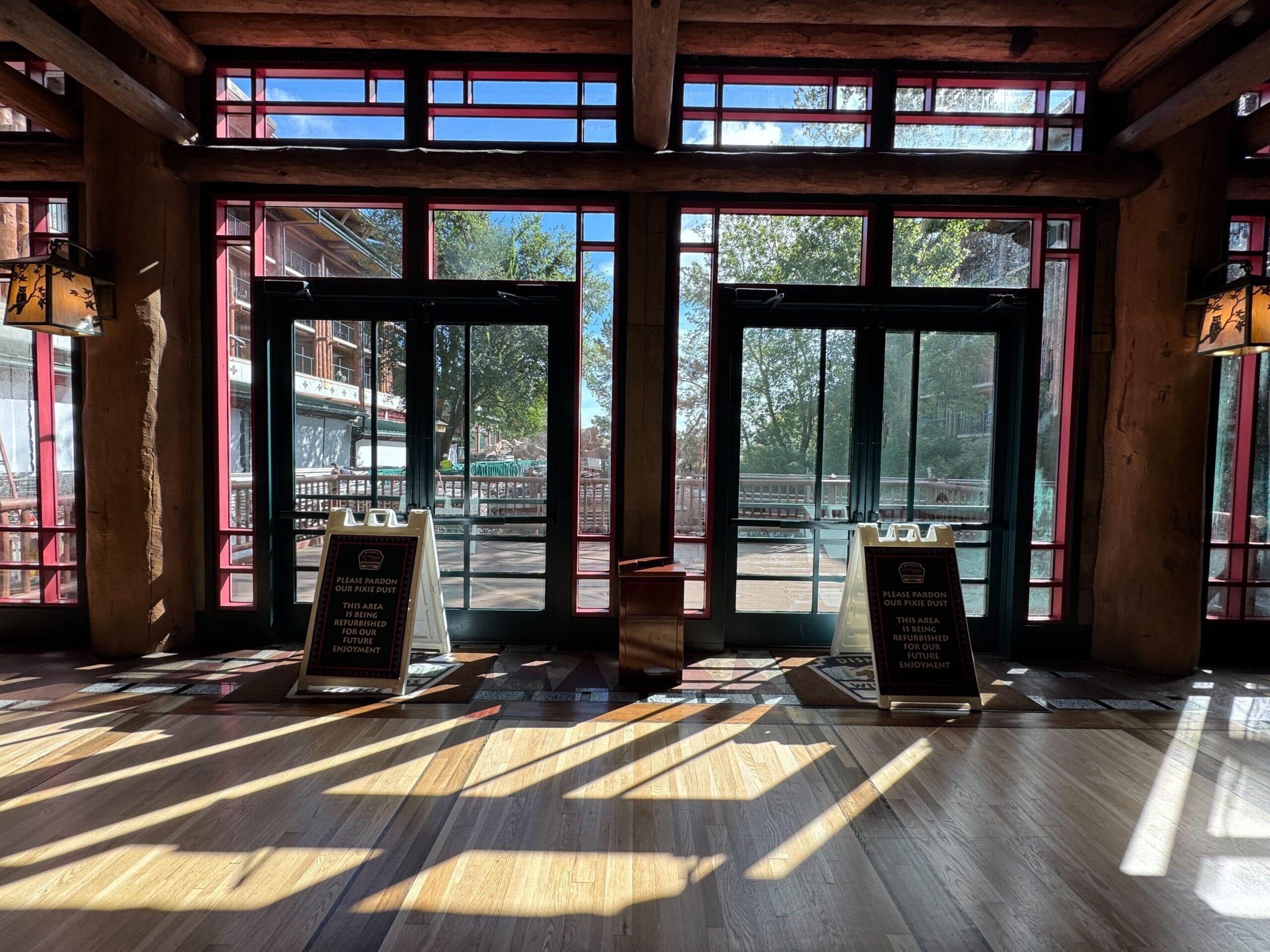 Sunlit lobby with large glass doors, wood beams, and two caution signs on the floor by the entrances.