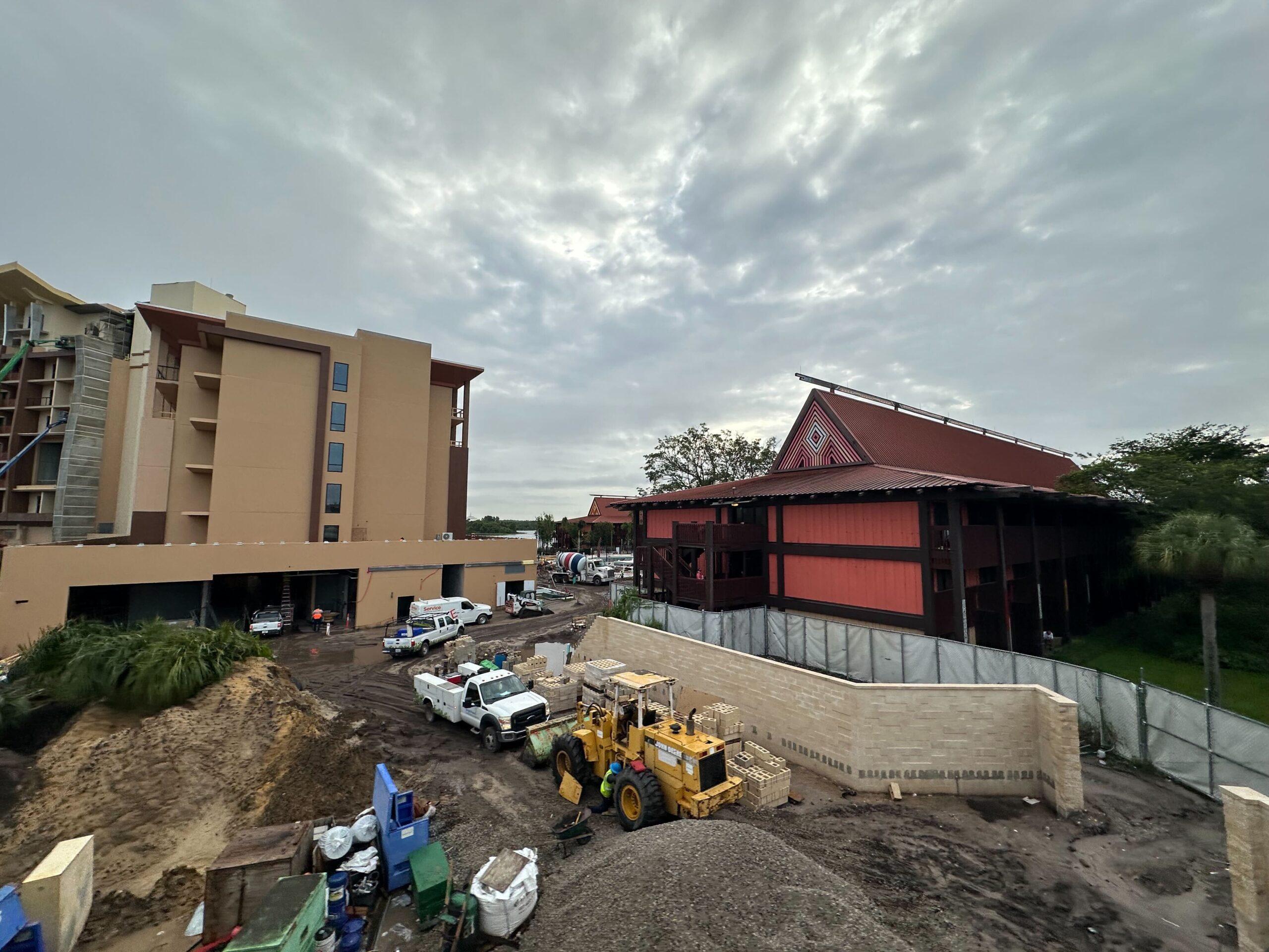 Construction site with heavy machinery, materials, and several workers beside a multi-story building and a smaller structure with a red roof on a cloudy day.
