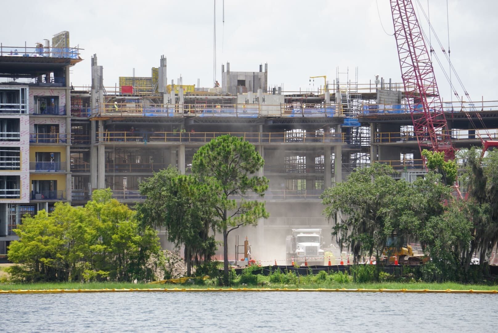 A construction site with a large building, trees in front, and a truck kicking up dust near a body of water.