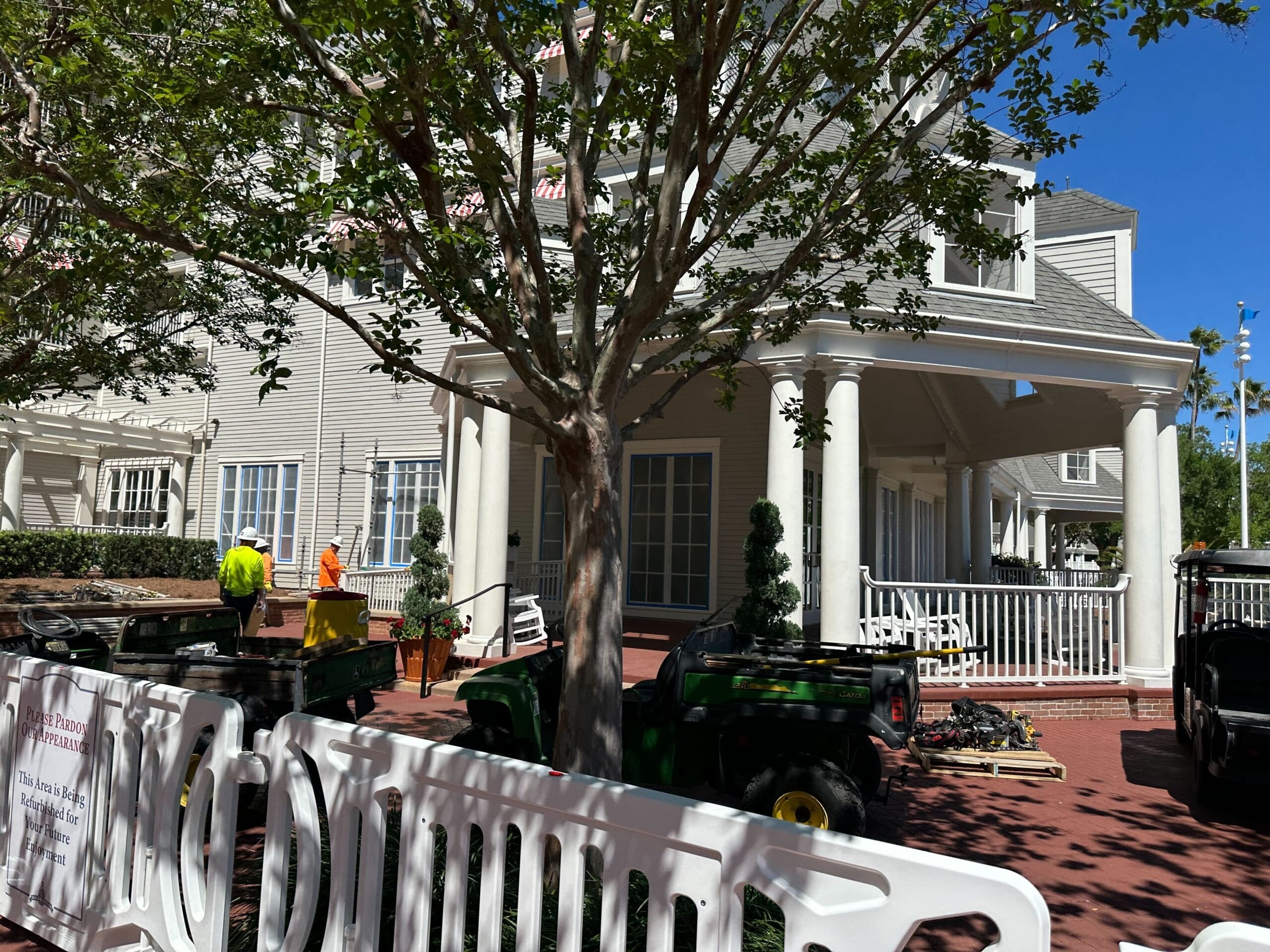 A two-story house with columns and a porch, reminiscent of Disney’s Yacht Club, is surrounded by trees. Construction workers and equipment are busy in front, with a white barricade adding to the scene's dynamic energy.